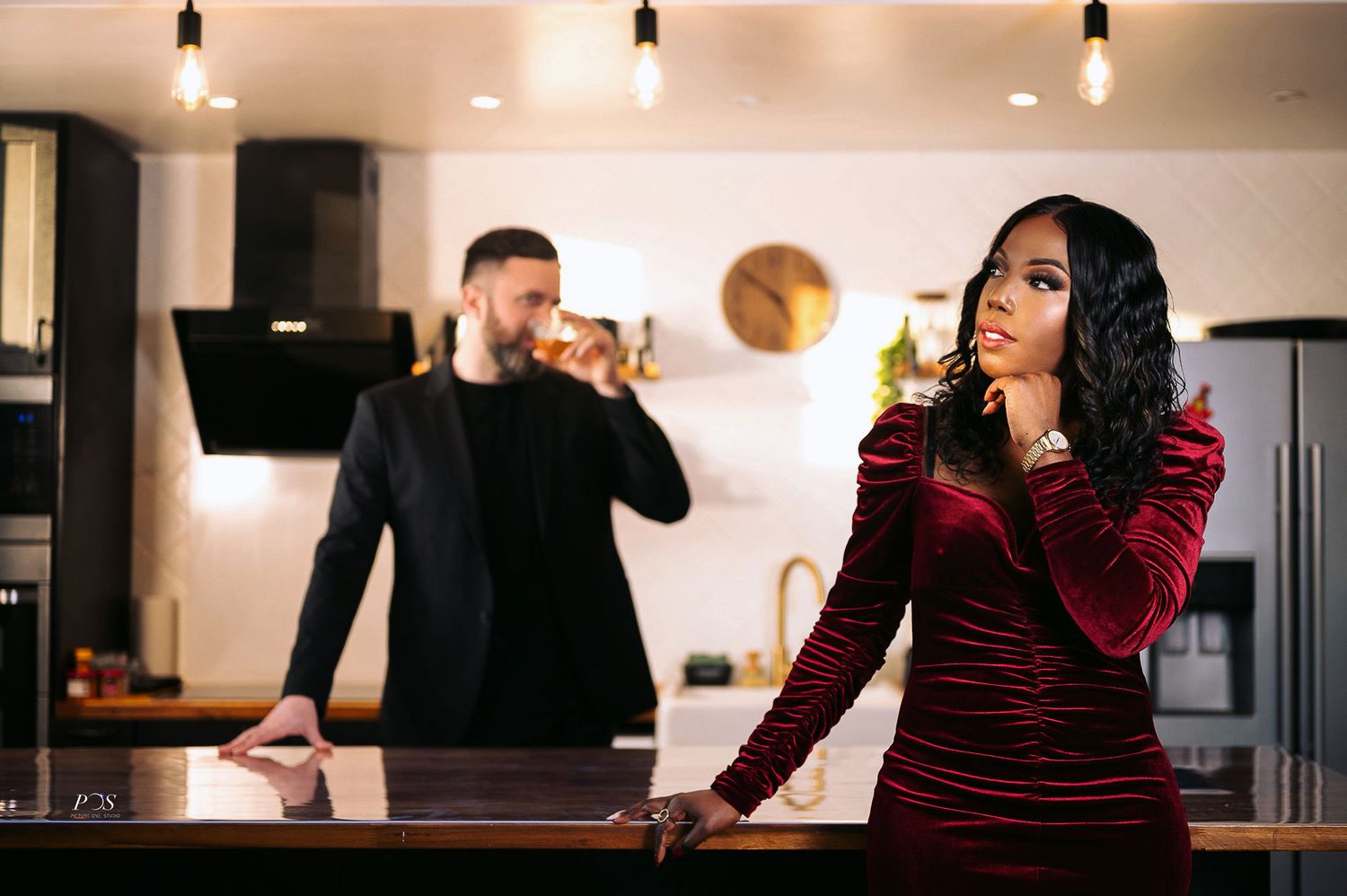 Couple standing apart in a stylish kitchen, woman in red velvet dress and man in black suit drinking.