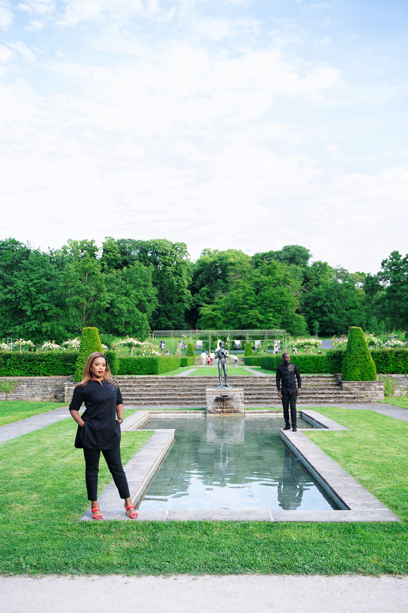 Couple dressed in black standing beside a rectangular garden fountain with lush green landscape.