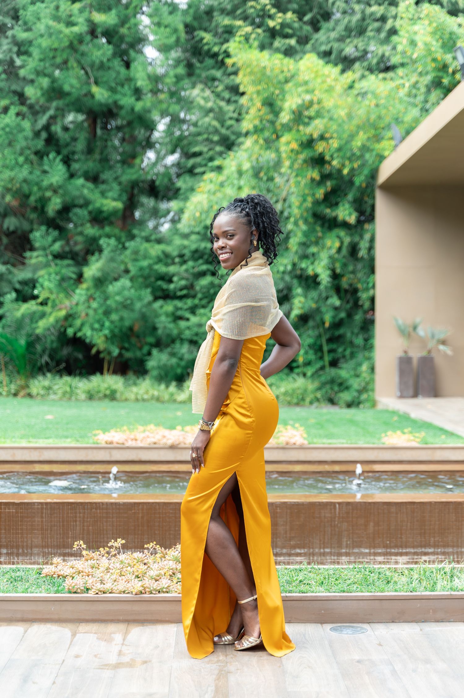 Woman in mustard yellow slit gown with sheer top poses gracefully by garden fountain at event.