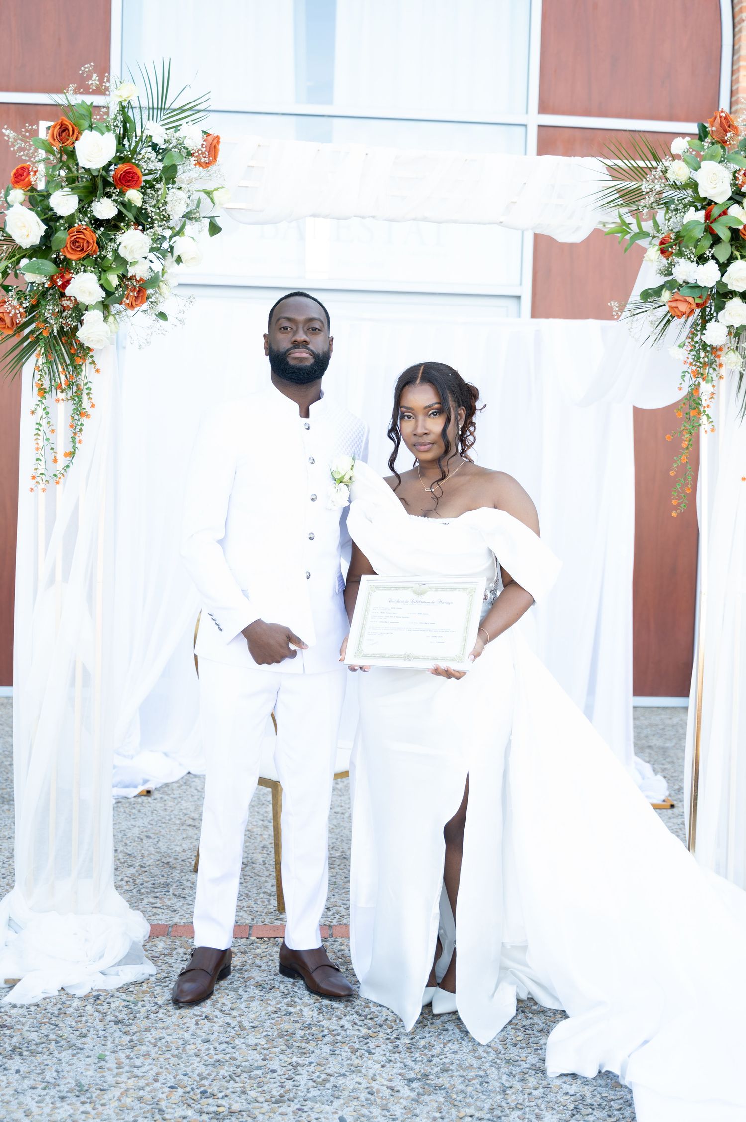 Bride and groom in white wedding attire holding marriage certificate under floral arch with orange and white flowers.