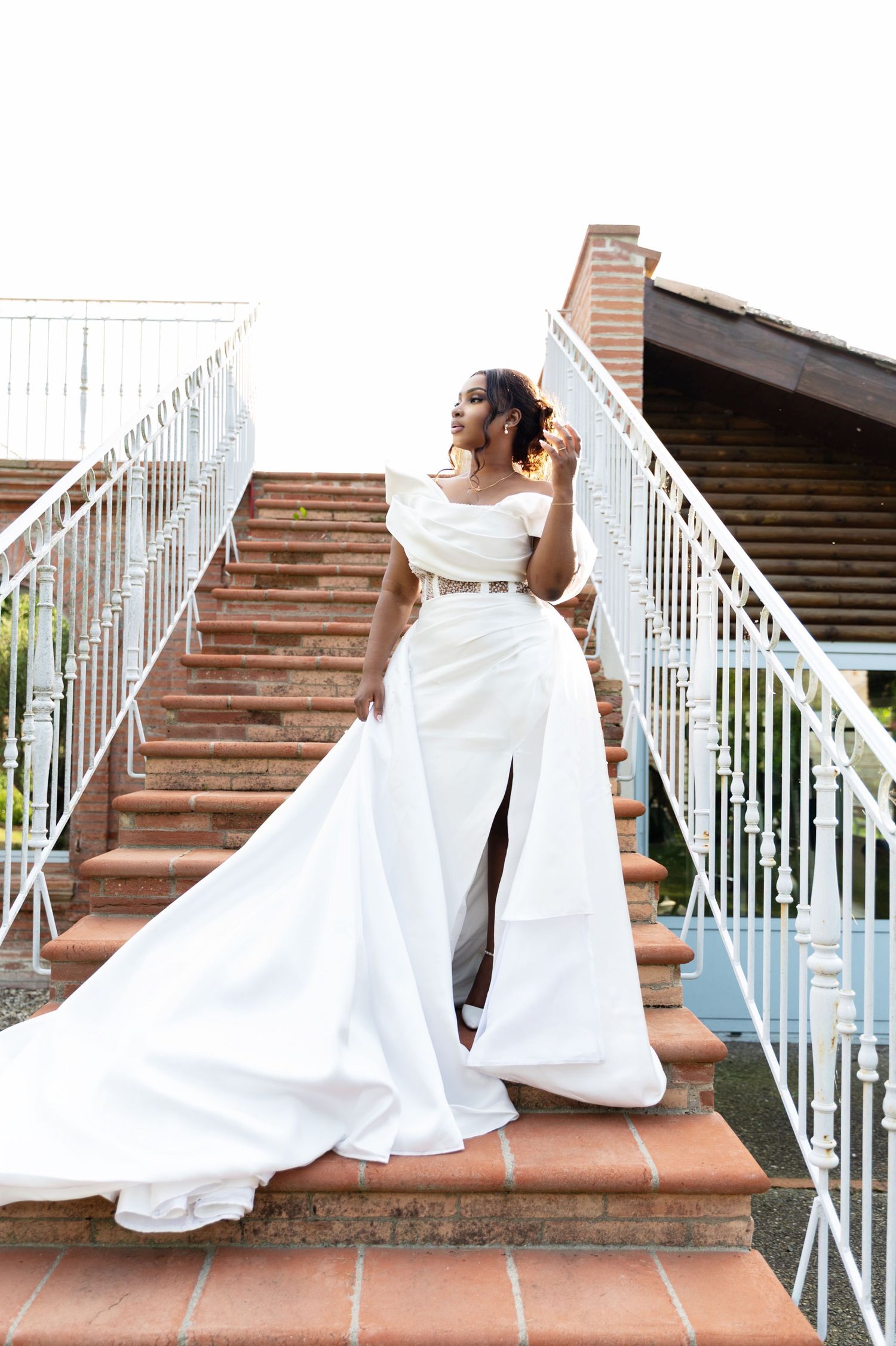 Bride in elegant white off-shoulder gown with dramatic train poses on brick staircase at outdoor wedding venue.