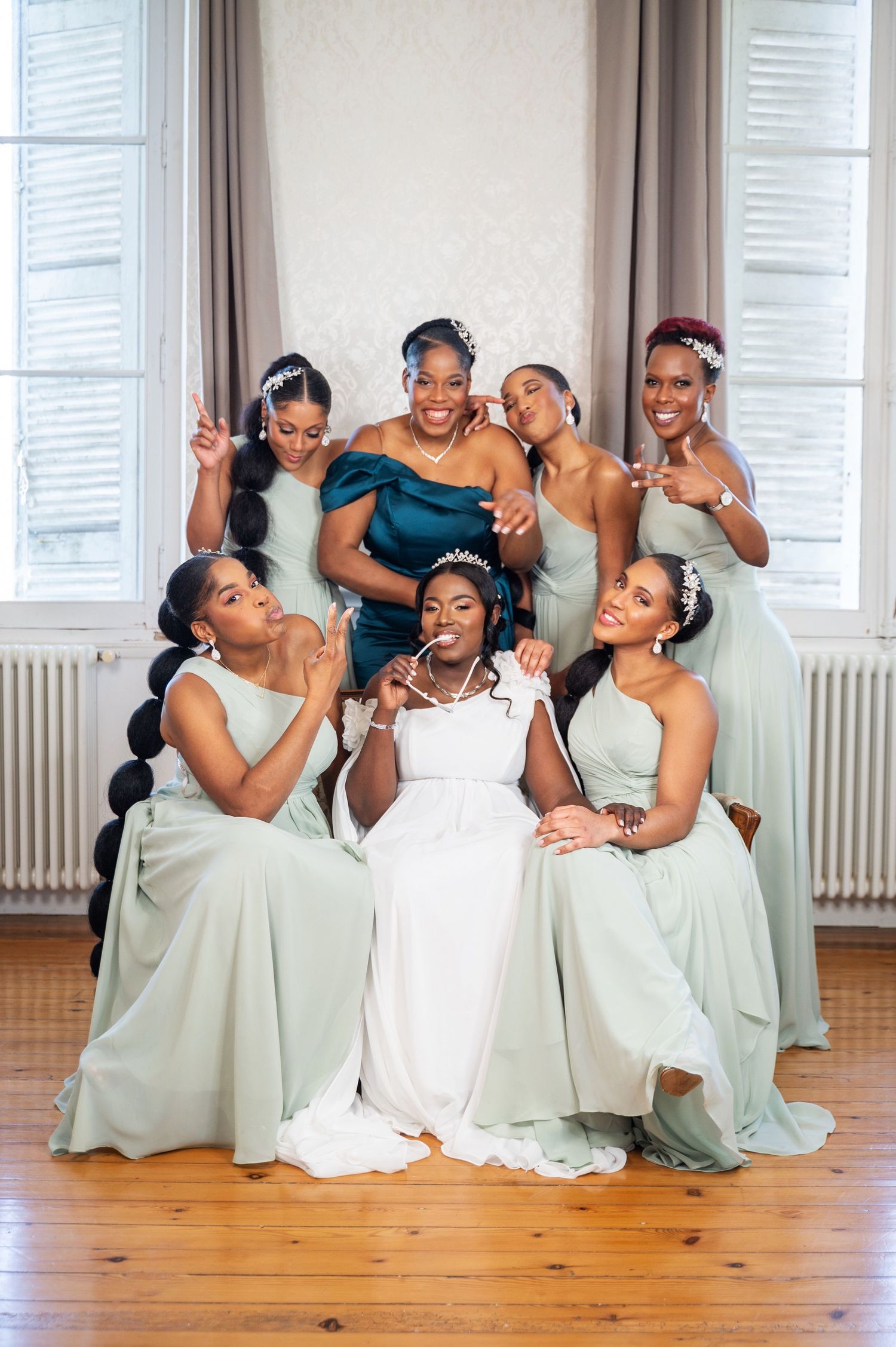 Bride seated in white gown surrounded by six bridesmaids in sage green dresses posing playfully indoors.