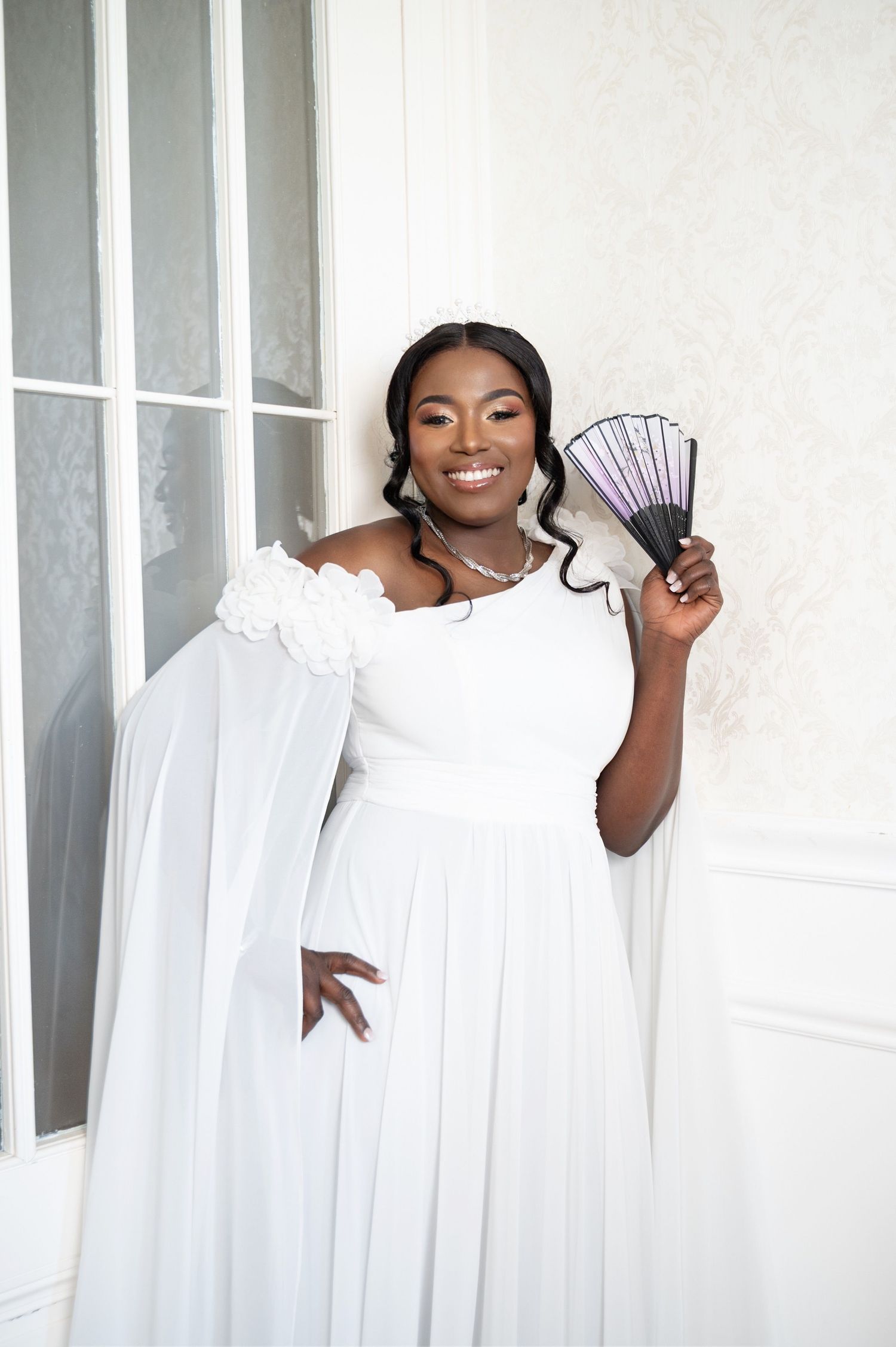 A smiling Black bride in a white off-shoulder gown holds a decorative fan while posing elegantly in a bright white room.