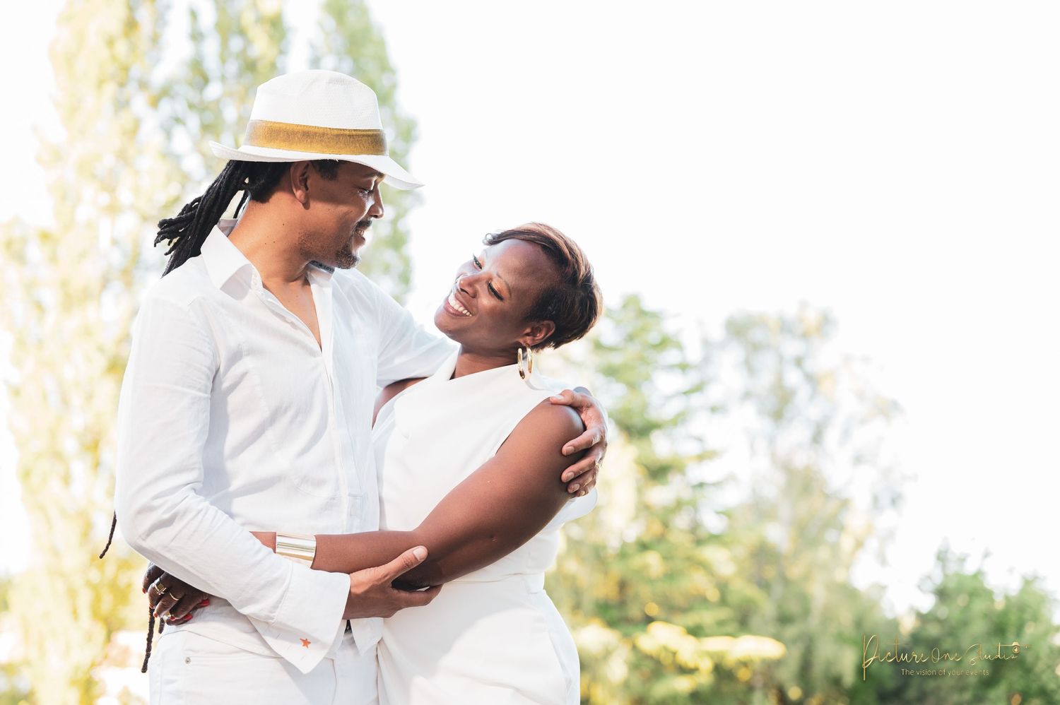 Couple in matching white outfits embracing and gazing at each other in sunlit outdoor setting.
