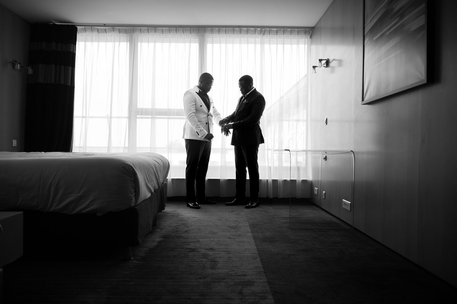 Two men in formal suits share a moment in a dimly lit hotel room before a wedding ceremony.