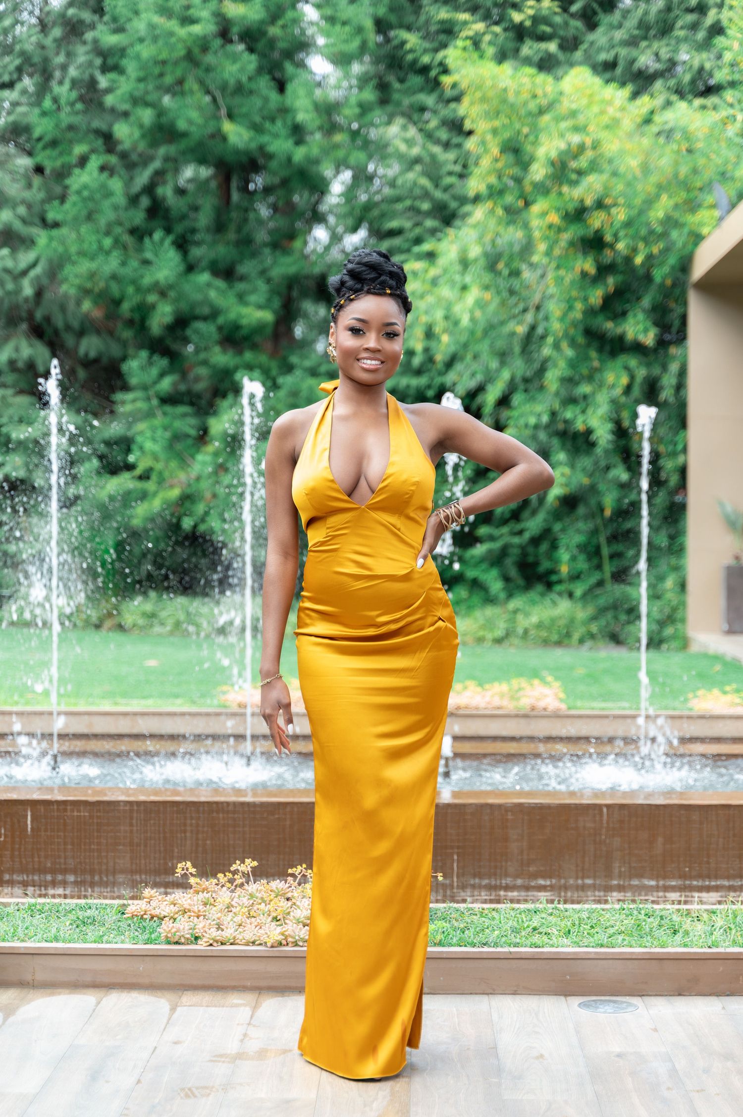 Smiling woman in mustard yellow halter gown posing confidently by garden fountain.