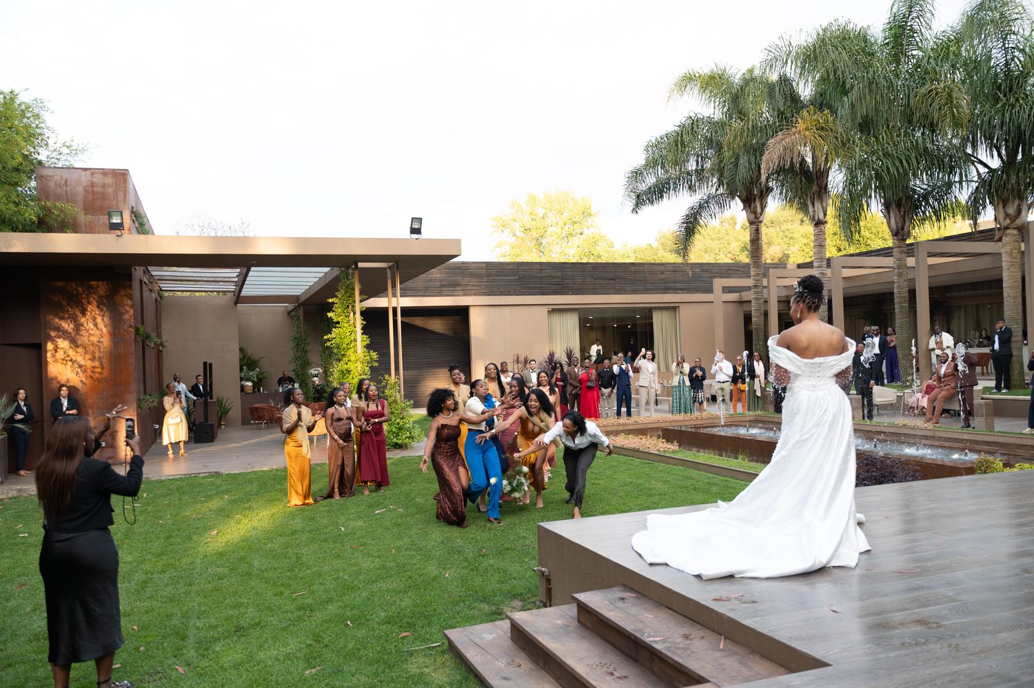 Bride in white gown descends steps toward wedding guests gathered on lush green lawn at outdoor venue.
