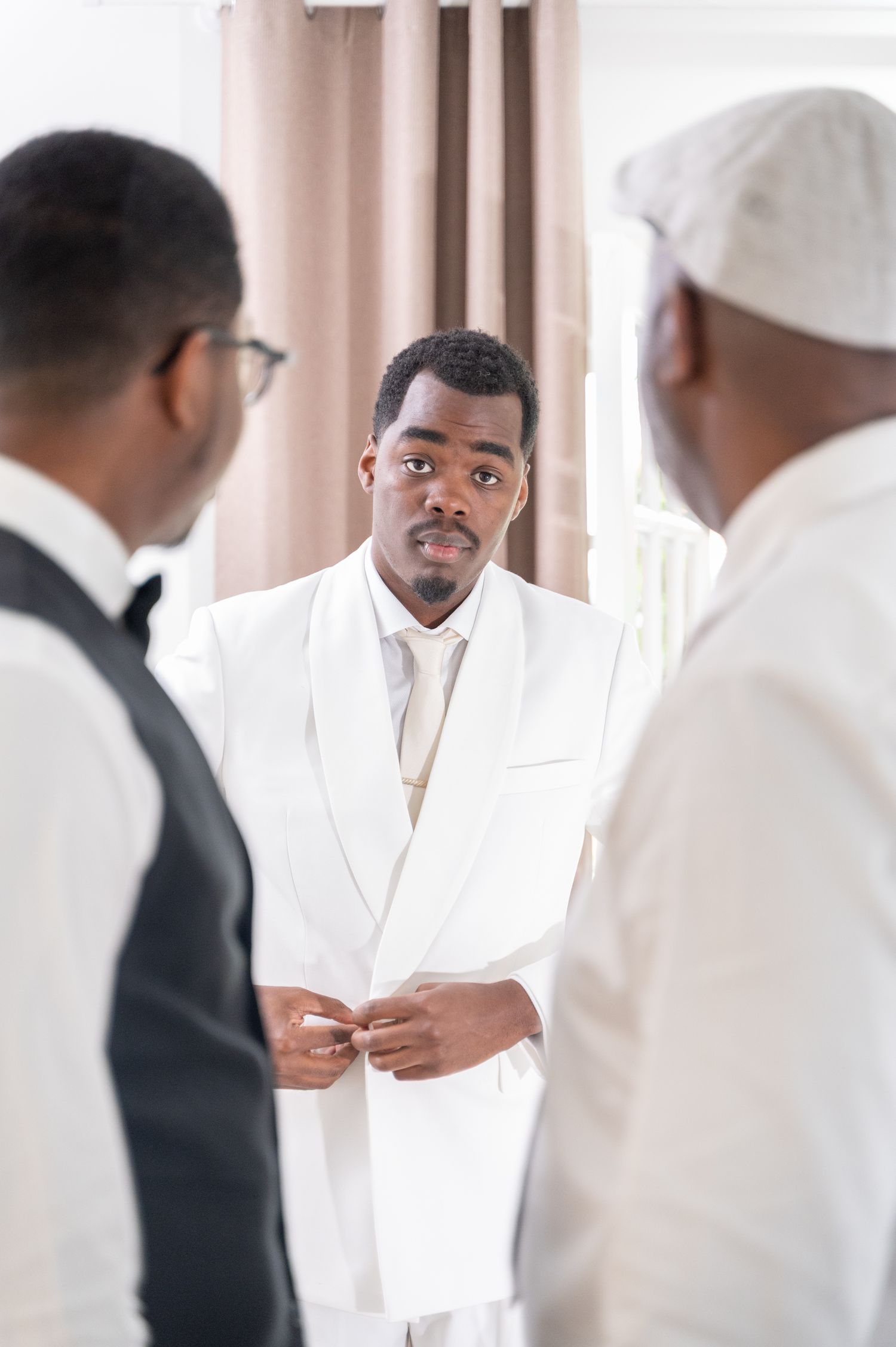 Groom in white tuxedo buttoning jacket while groomsmen assist during wedding preparations.