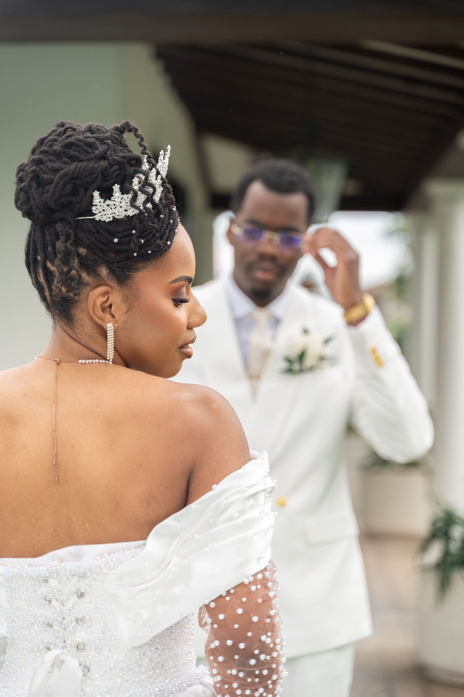 Bride in pearl off-shoulder wedding dress with crystal tiara while groom adjusts glasses behind her.