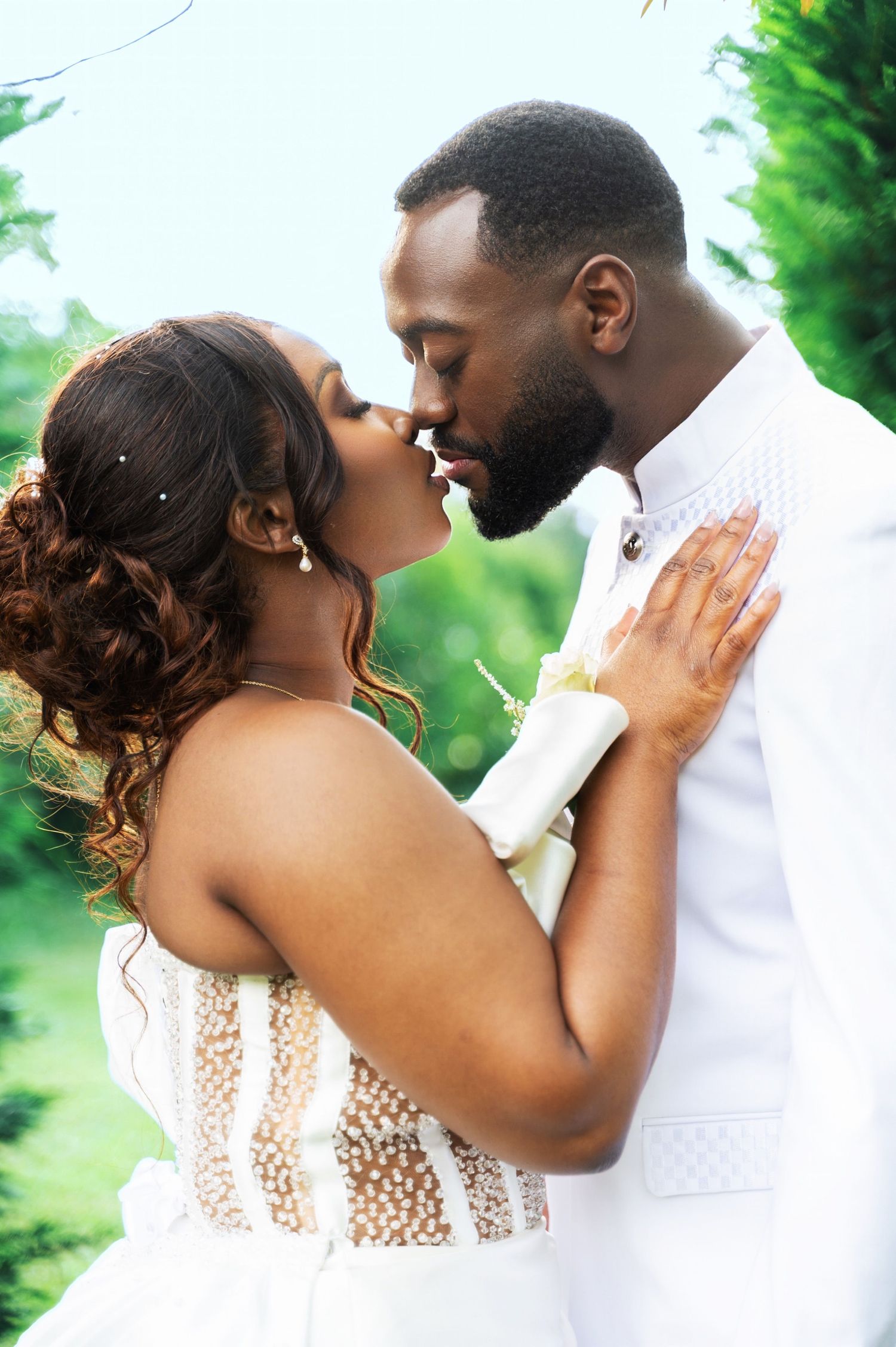 A Black bride and groom share a romantic kiss outdoors, both dressed in elegant white wedding attire.