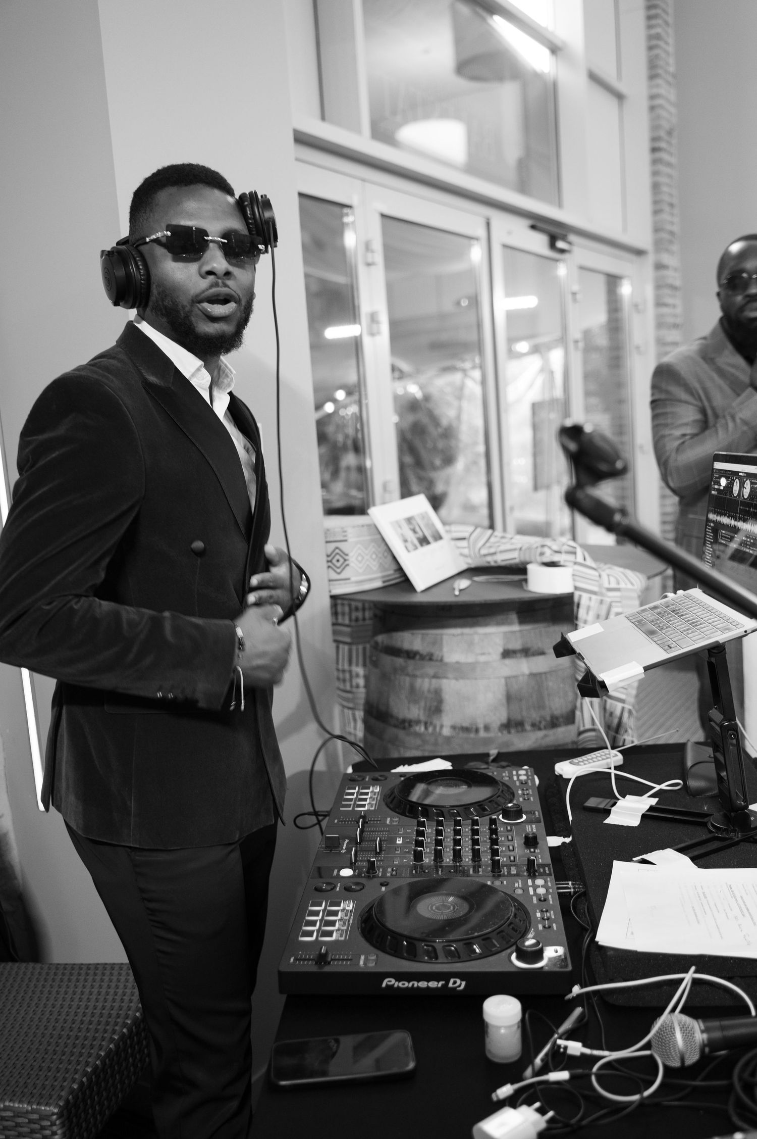 Black and white photo of stylish DJ with headphones and sunglasses operating Pioneer DJ equipment at event.