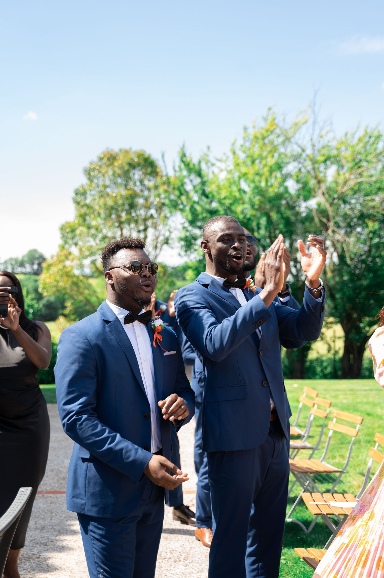 Two groomsmen in navy suits with bow ties clapping and celebrating at outdoor wedding ceremony.