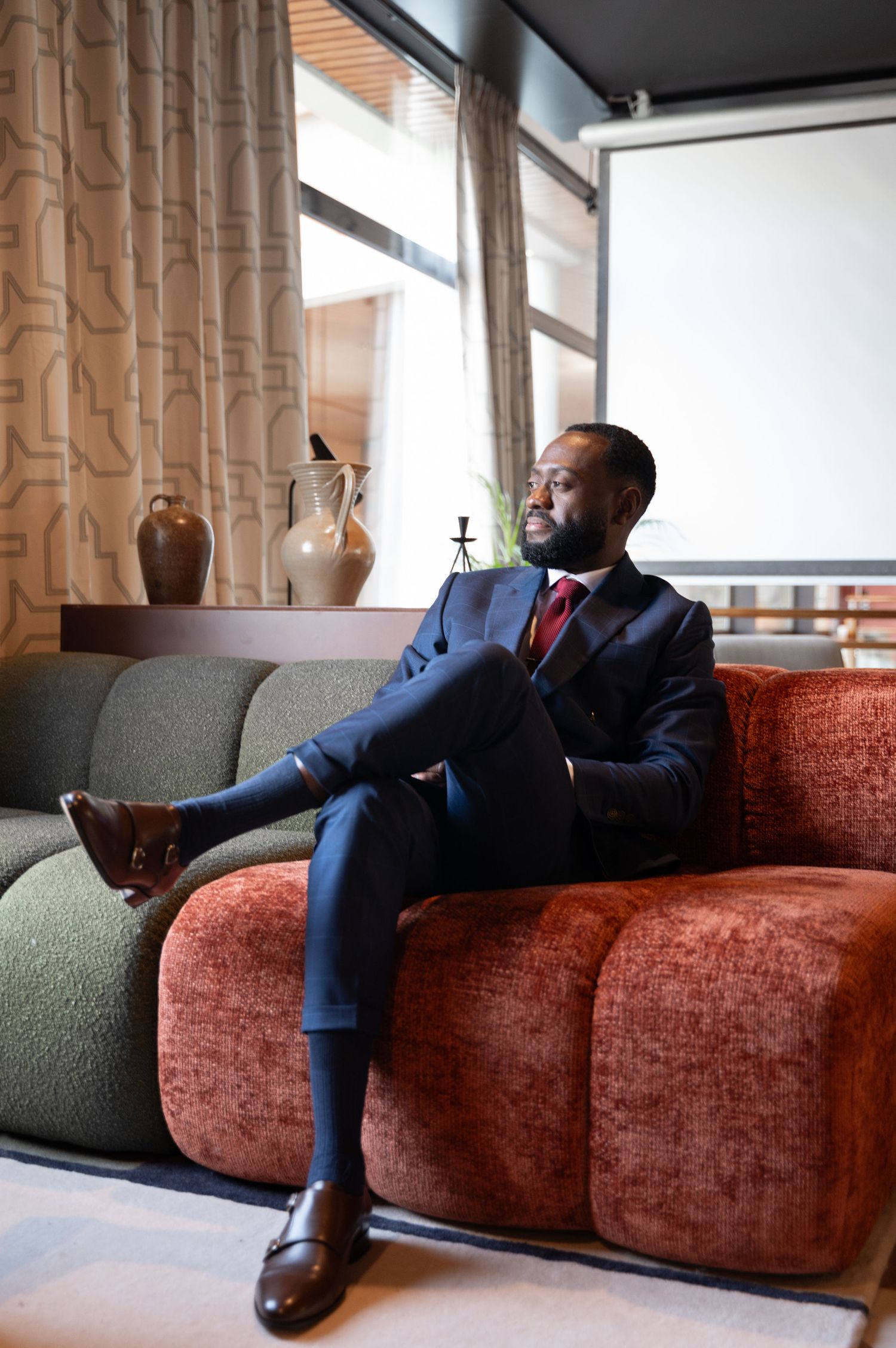 Confident Black man in navy suit and red tie sitting cross-legged on a colorful velvet sofa by large windows.