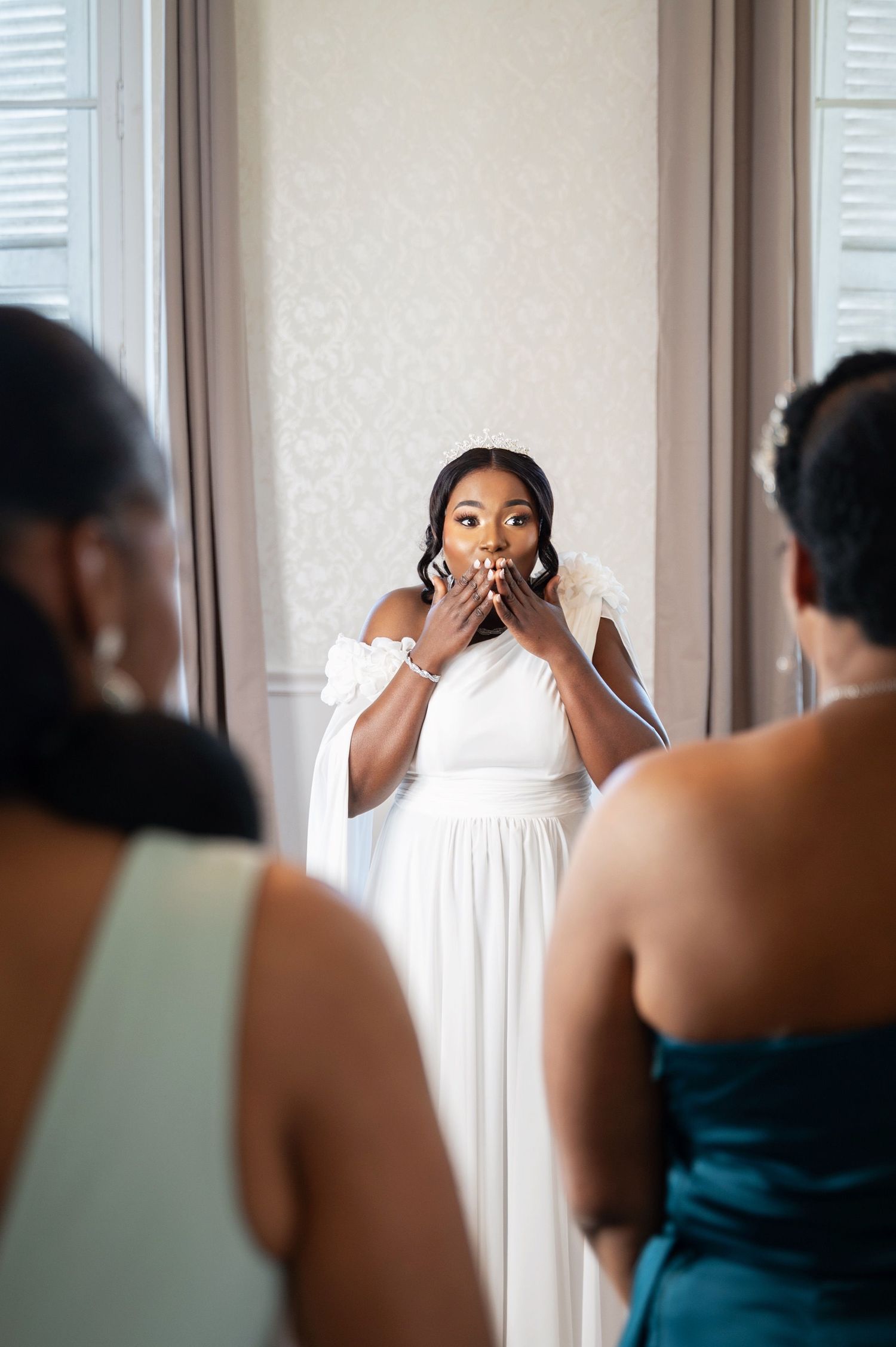 A surprised Black bride in a white off-shoulder gown covers her mouth with both hands during an emotional first look reveal.