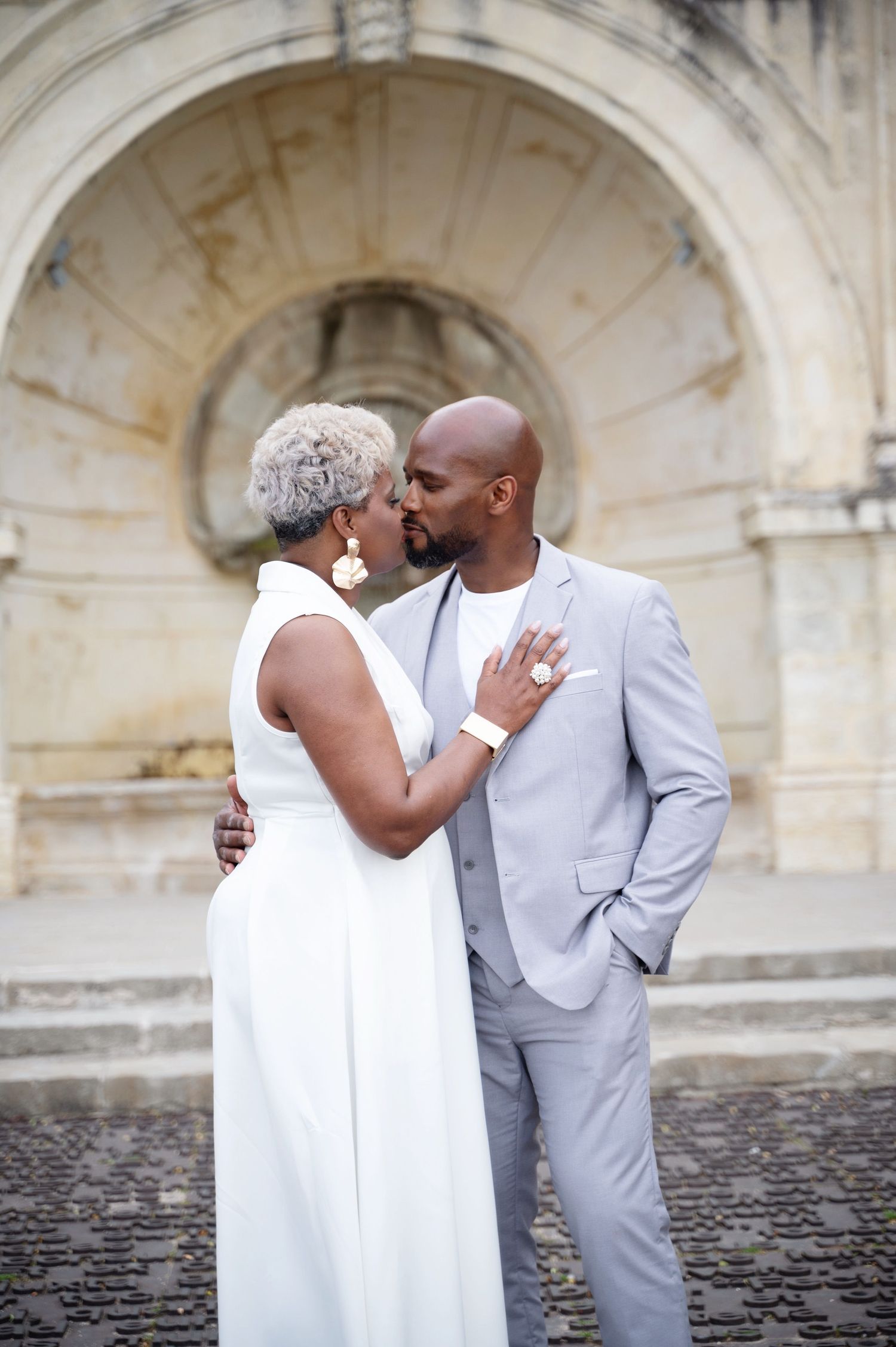 Couple sharing a kiss in front of a grand stone archway on cobblestone ground.