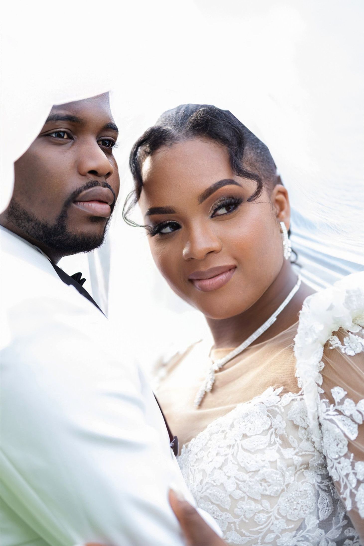 Close-up portrait of bride in lace gown and groom in white suit gazing at camera.