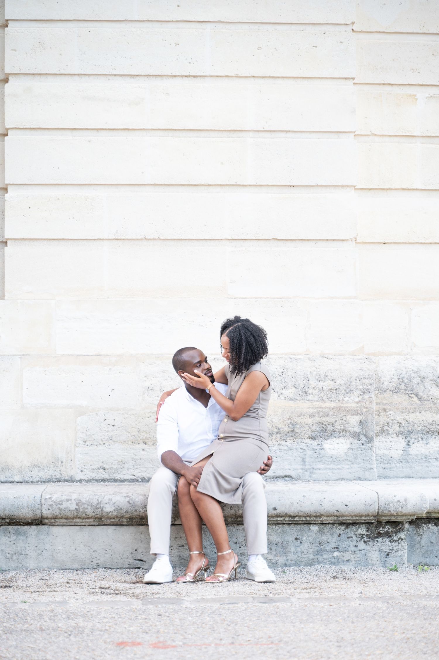 A couple sits intimately on stone steps against a white wall, the woman gently touching the mans face.