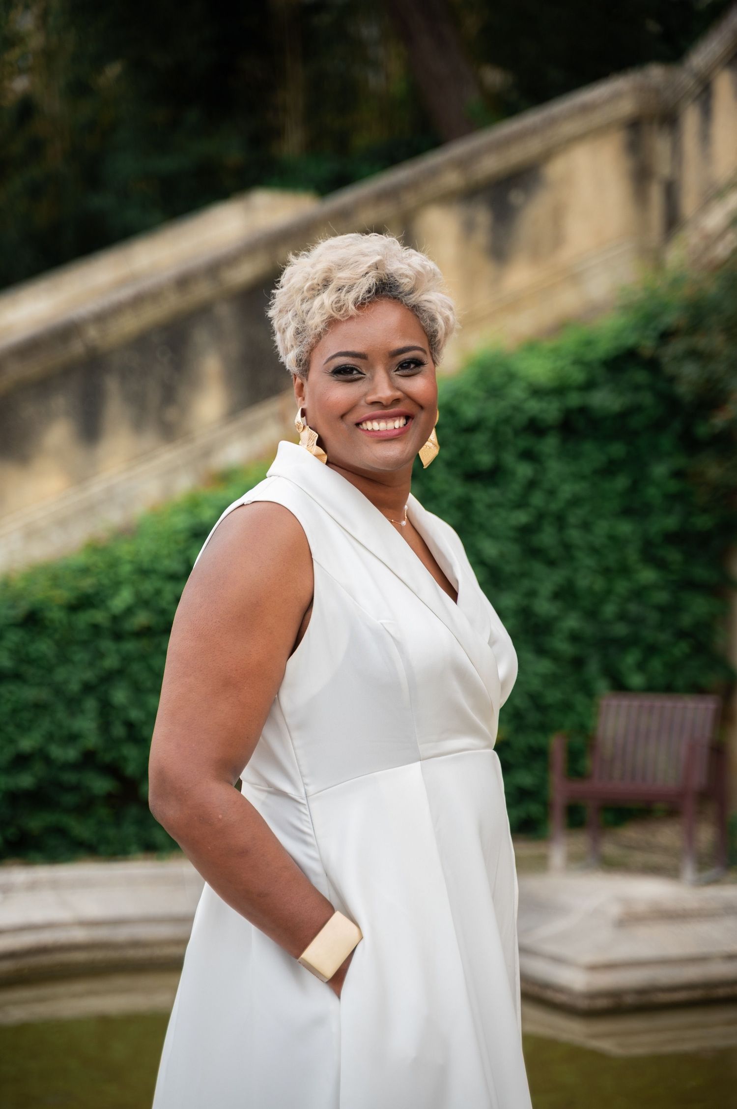 Smiling Black woman in white sleeveless dress with gold earrings posing in a lush garden setting.