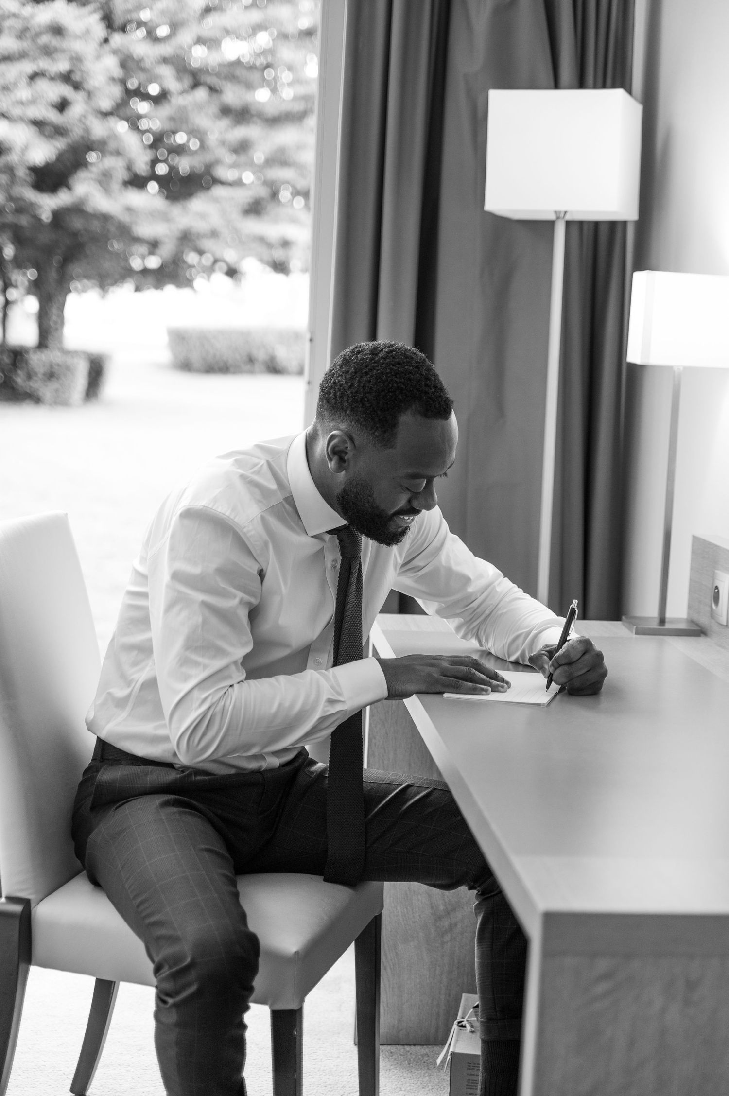 Groom in white shirt and tie writes a letter at a desk in black and white photo.