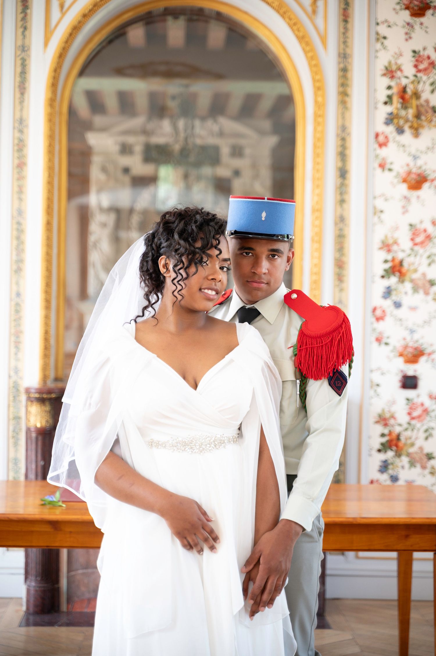 Black bride and military groom share tender moment inside ornate floral decorated hall.