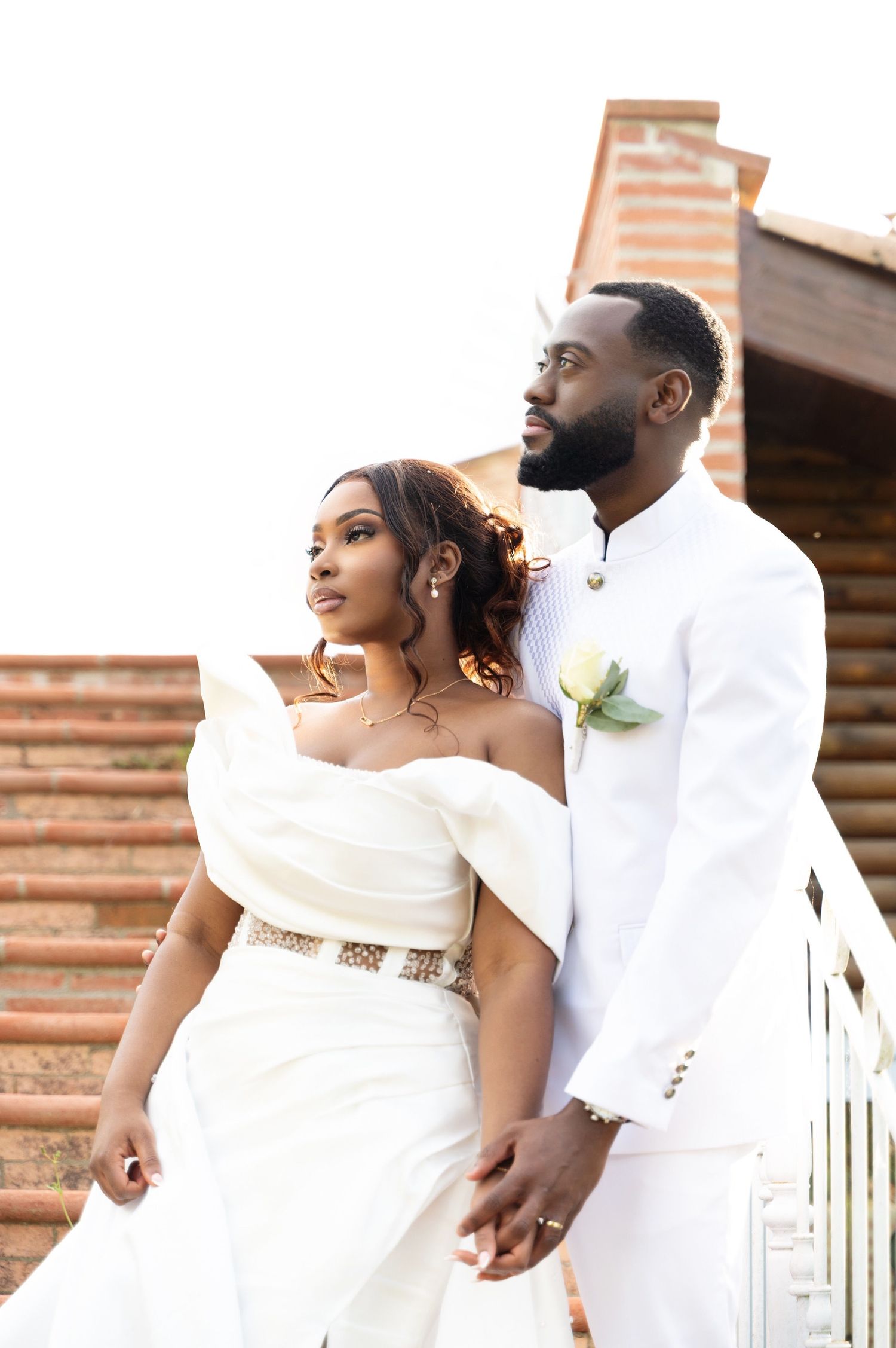 Elegant wedding couple in white attire standing together on brick stairs gazing into the distance.