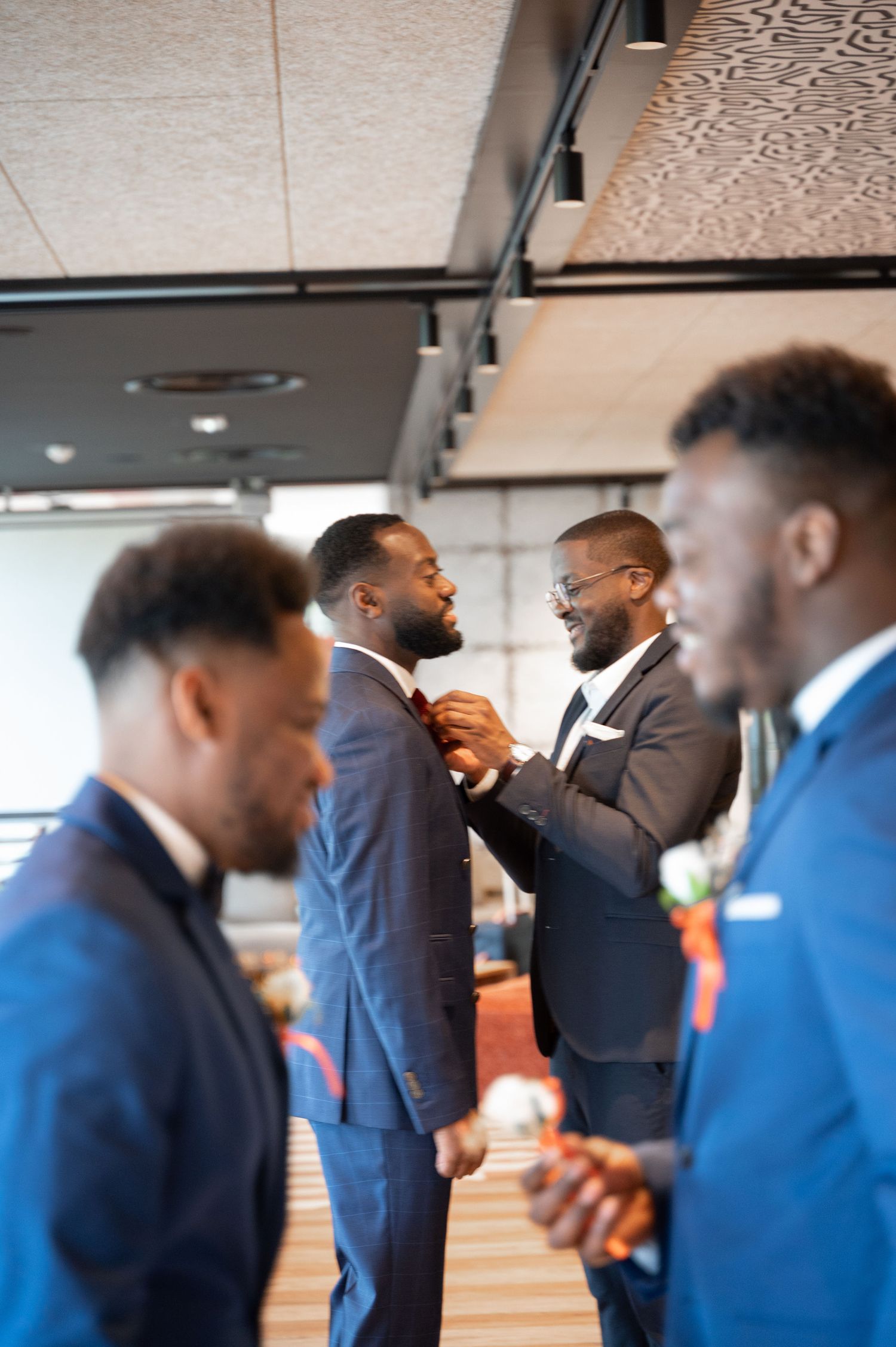Groomsman in dark suit adjusting another mans tie during wedding preparations in a modern venue.