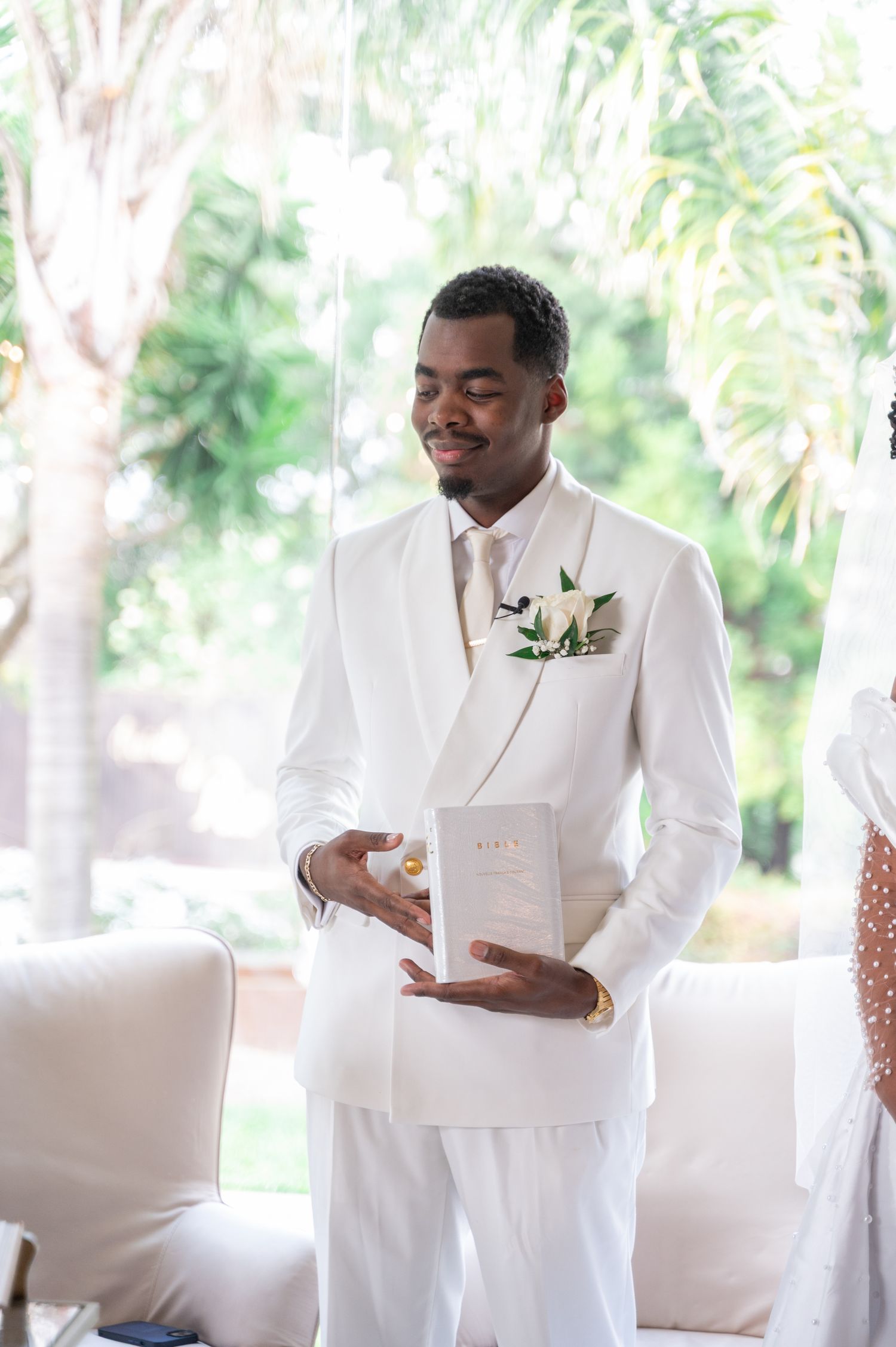 Groom in white suit holding Bible with floral boutonniere at wedding ceremony.