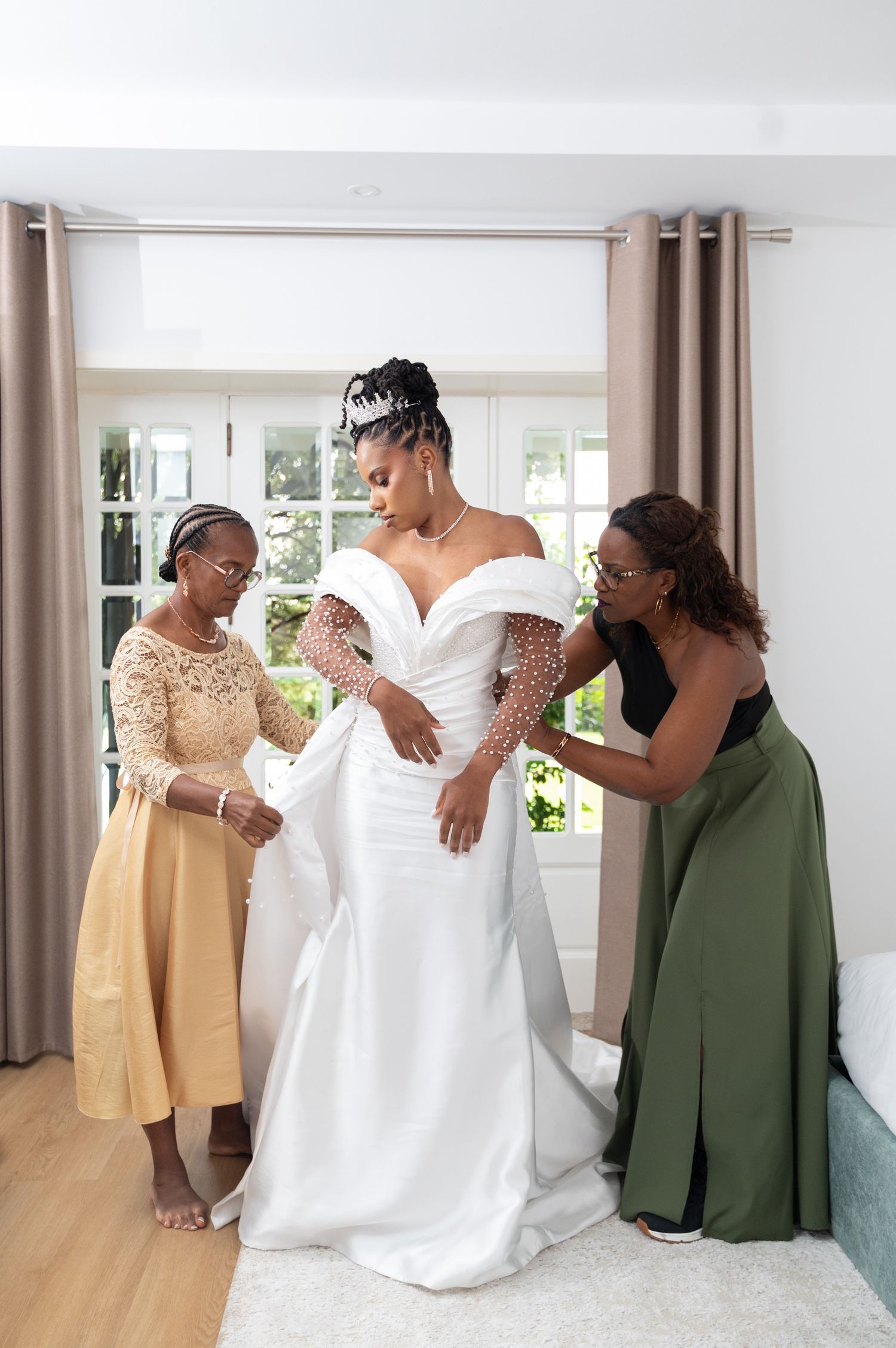 Black bride assisted into white wedding gown by two women in elegant attire.
