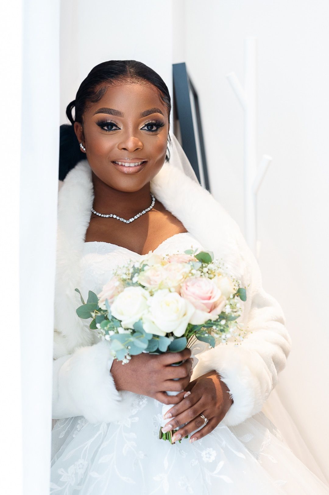 Smiling Black bride in white strapless gown, fur wrap, veil, and diamond necklace holding pastel bouquet.