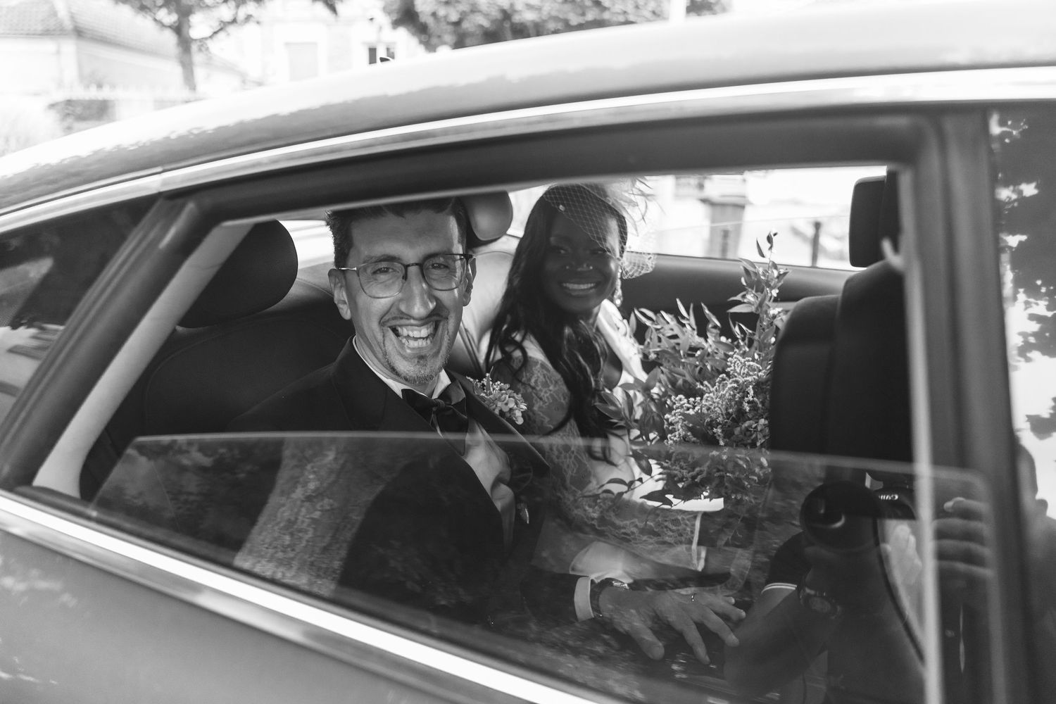 Happy newlywed couple smiles through car window in a joyful black and white wedding photo.