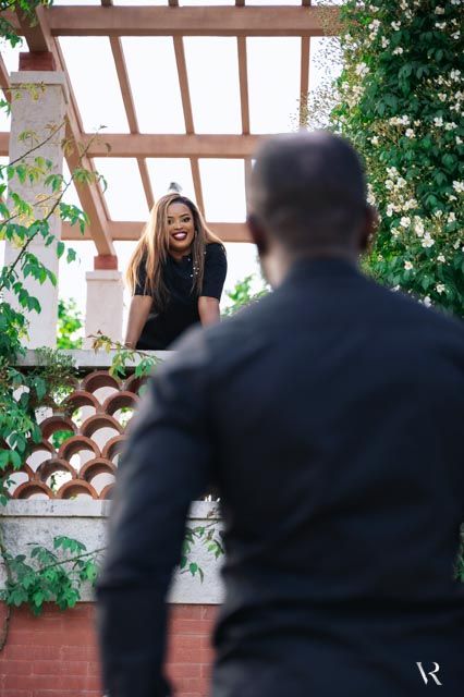 A smiling woman leans over a lattice wall under a pergola, looking down at a man in a black jacket outdoors.