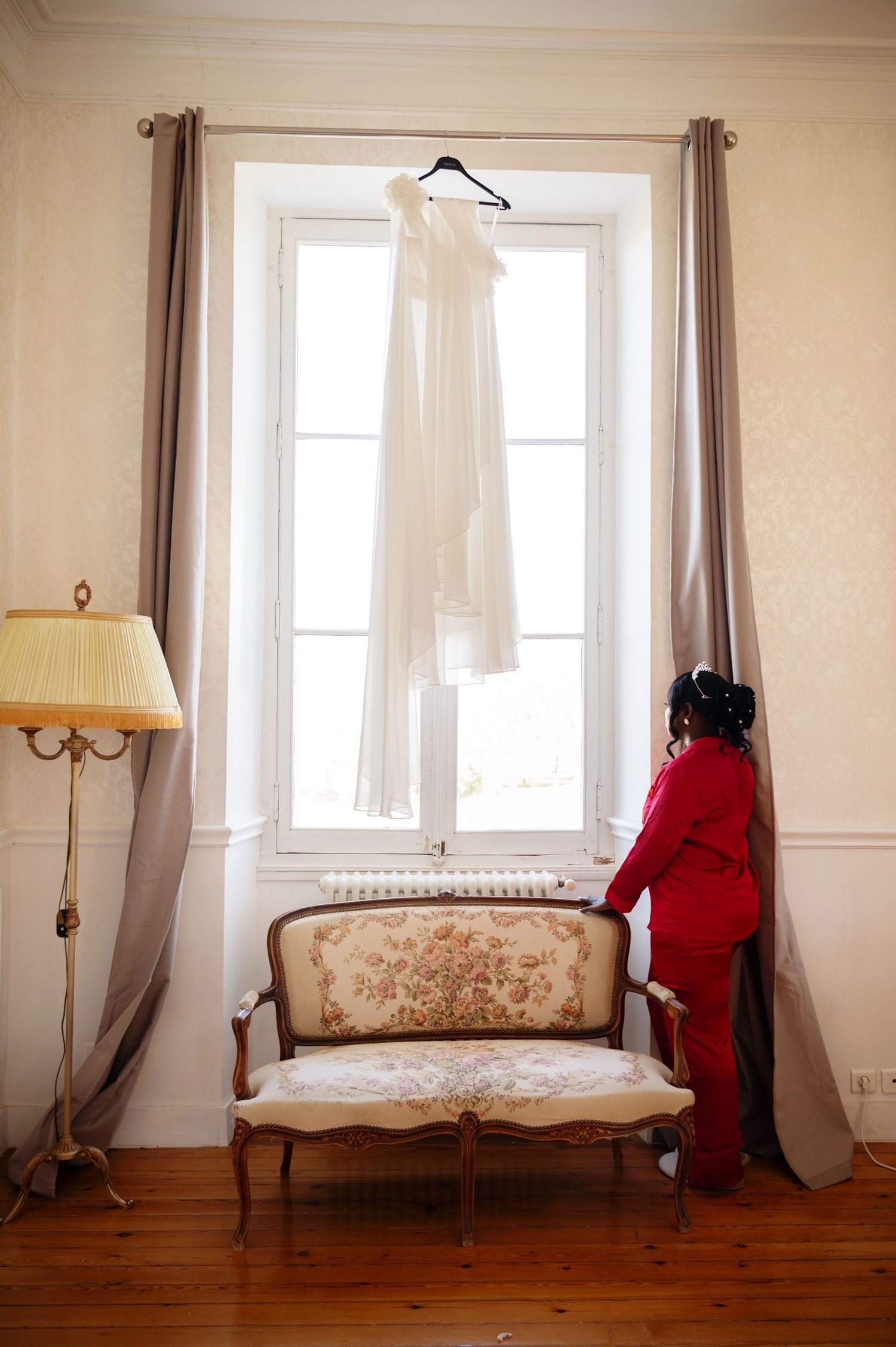 Bride in red robe admiring white wedding dress hanging on window curtain rod above vintage floral settee.