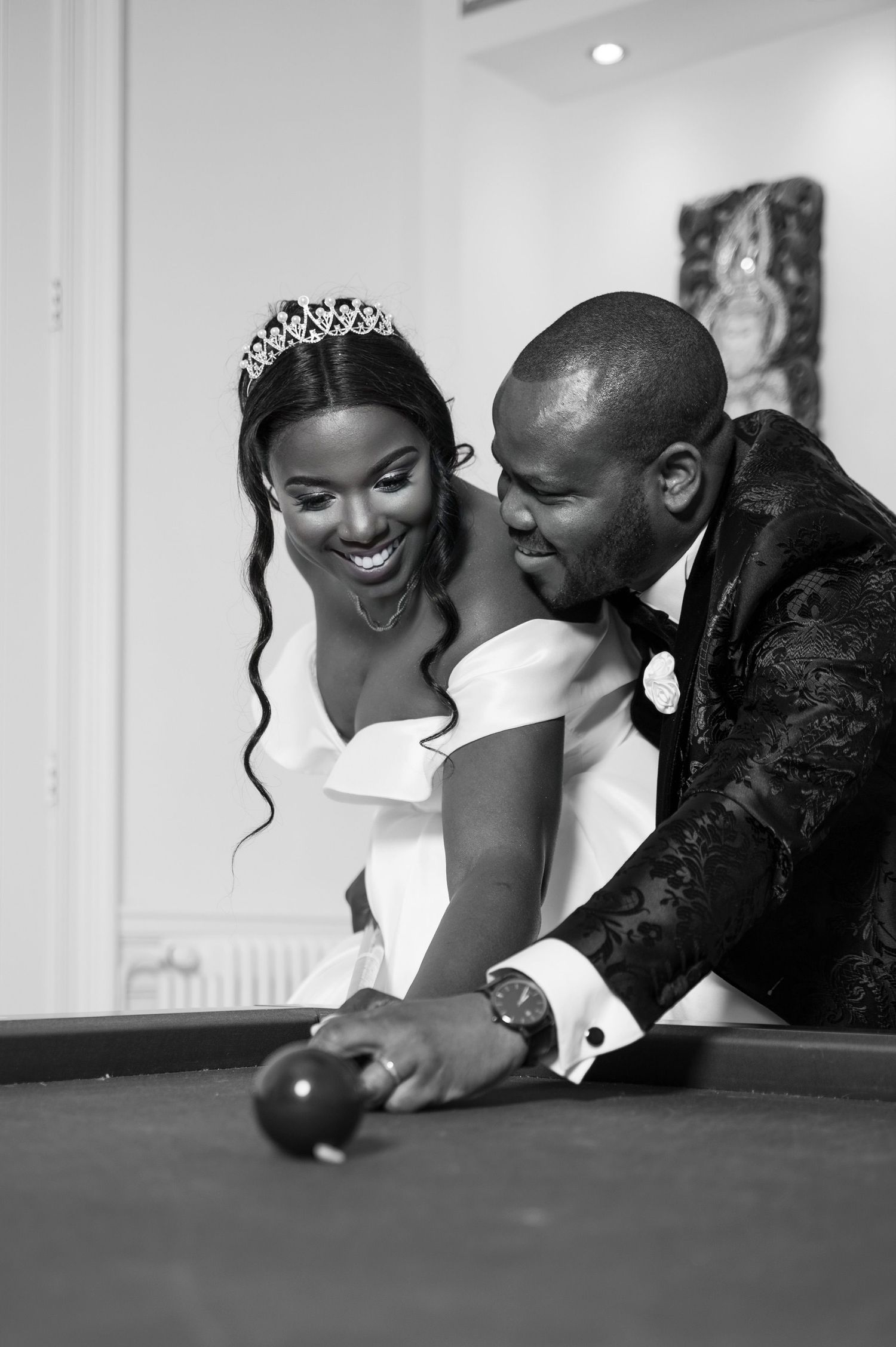 Black and white photo of a laughing Black bride and groom playing billiards together at their wedding.