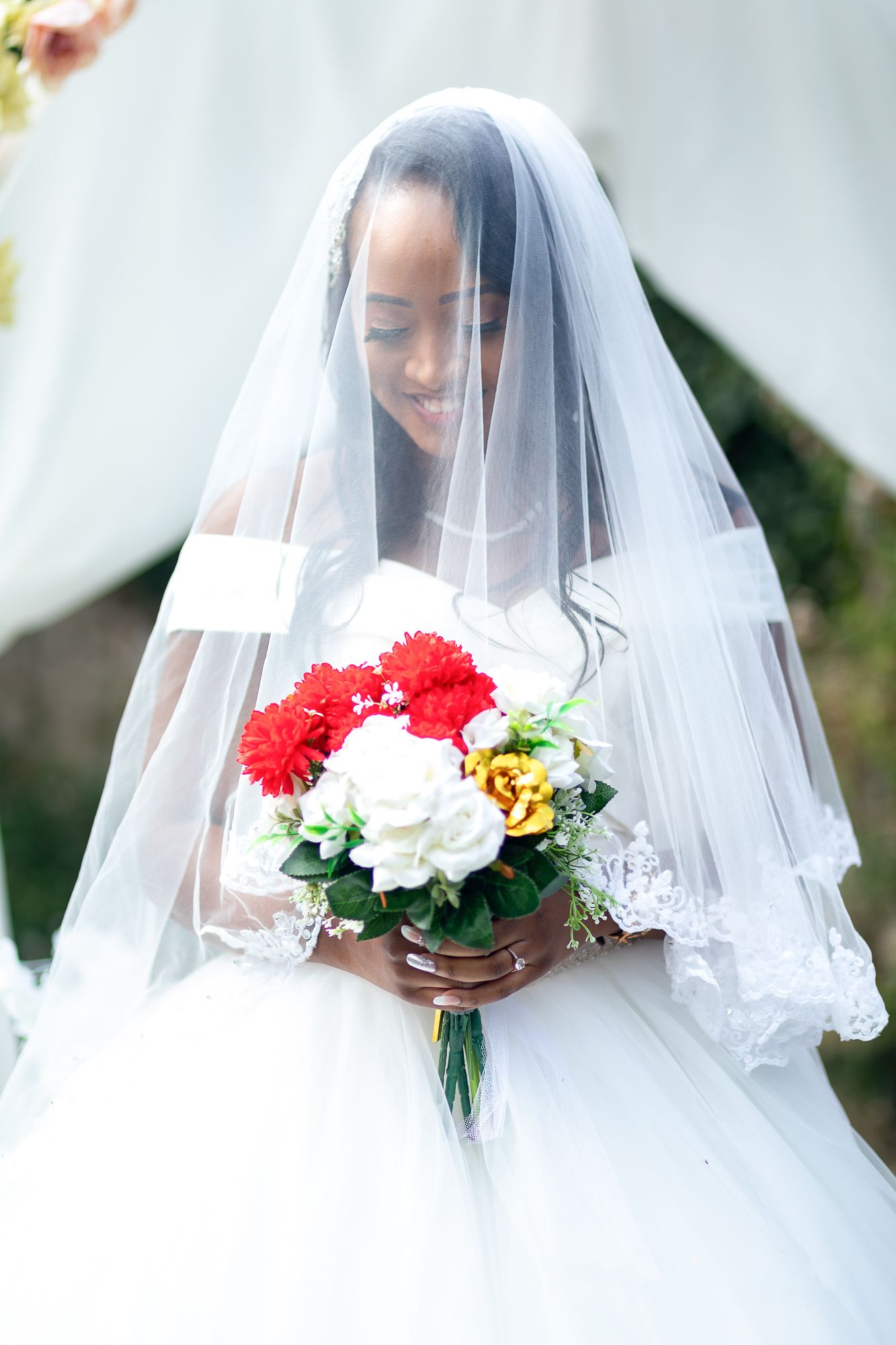 Beautiful African bride in white lace gown and veil holding red and white bouquet.
