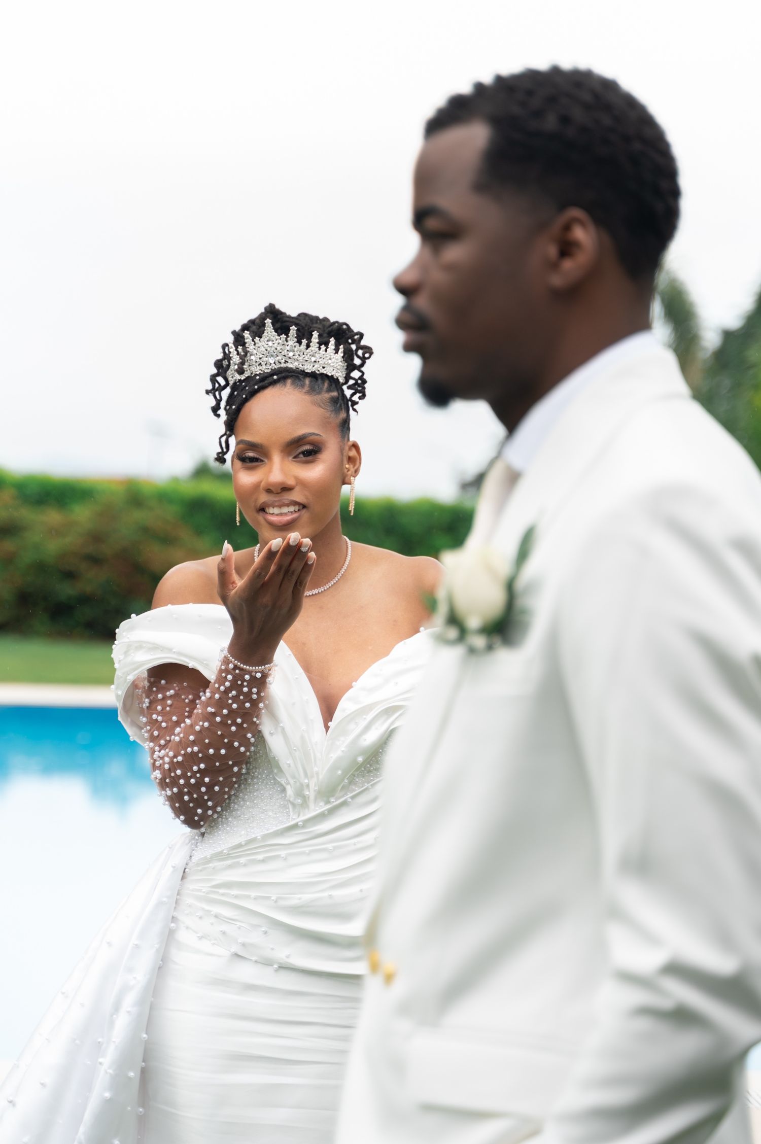 Bride in white beaded gown and tiara blowing a kiss to groom in white suit near a pool.