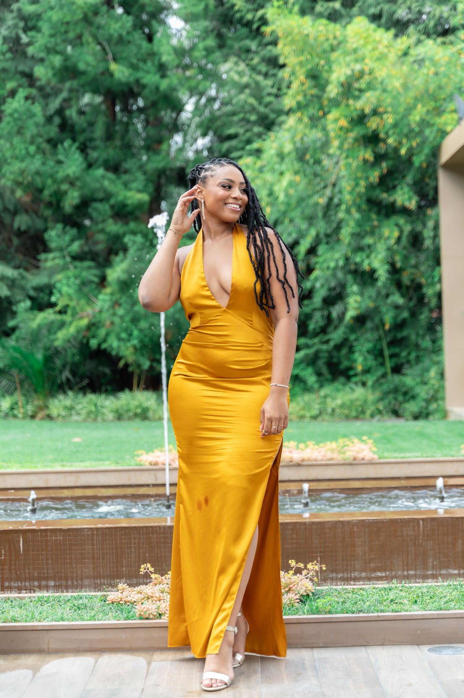 Smiling woman in mustard yellow halter gown with locs poses near garden fountain at outdoor event.
