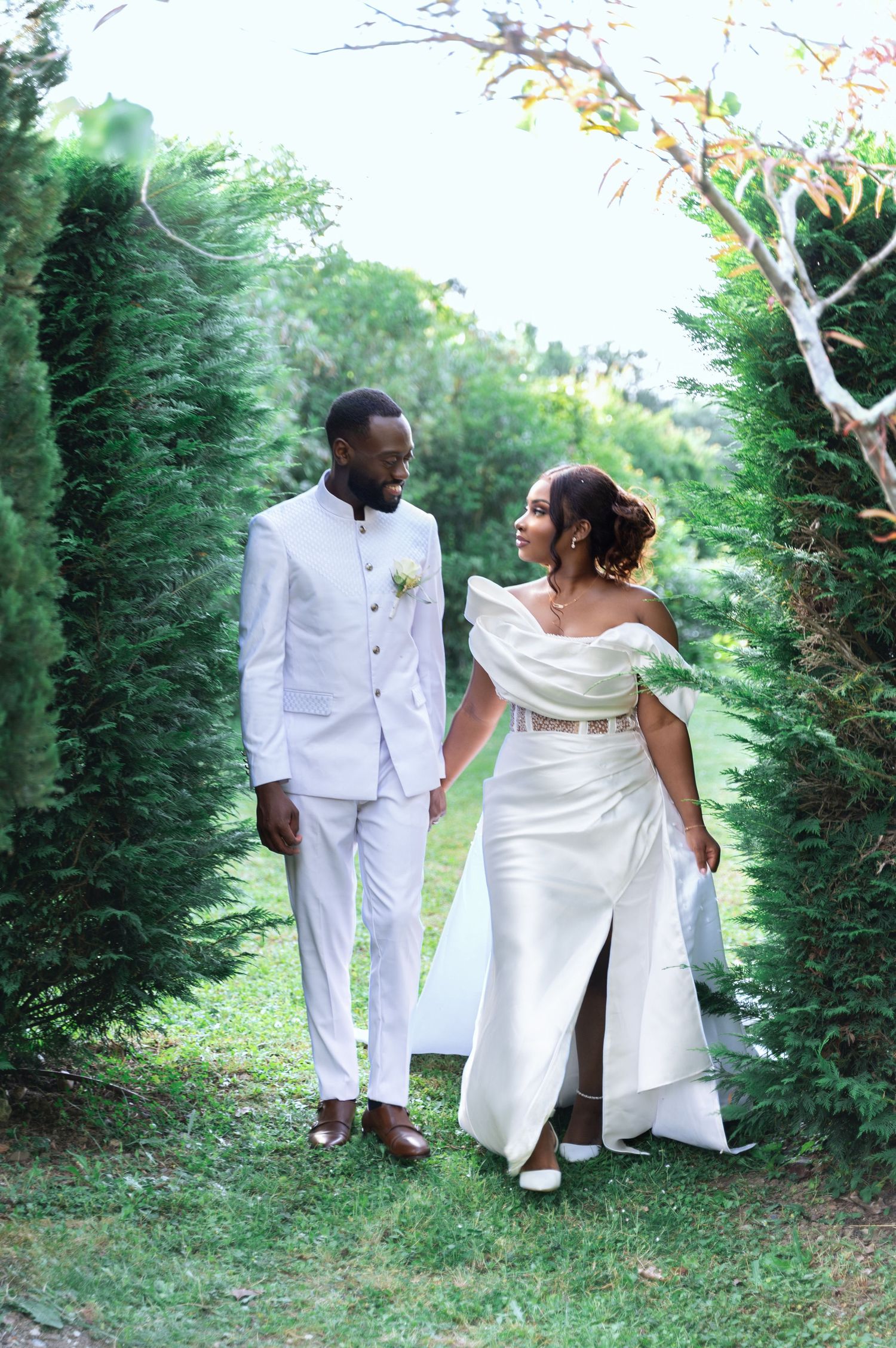 An elegant Black couple in white wedding attire hold hands while walking between tall green hedges.