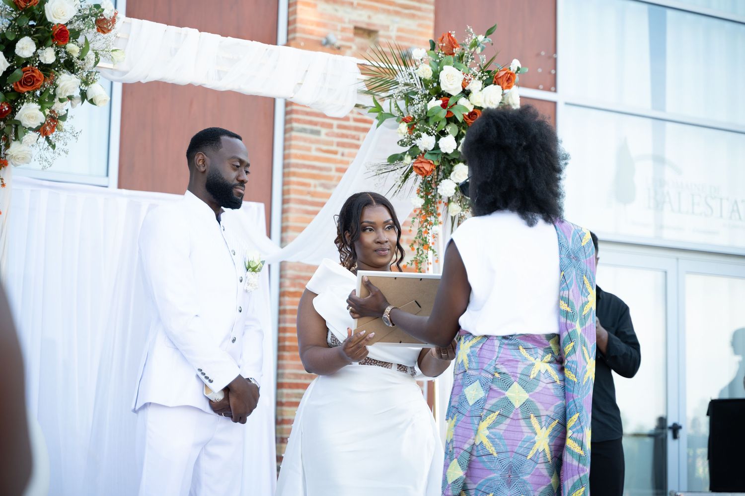Female officiant in colorful dress leads outdoor wedding ceremony for couple in white.