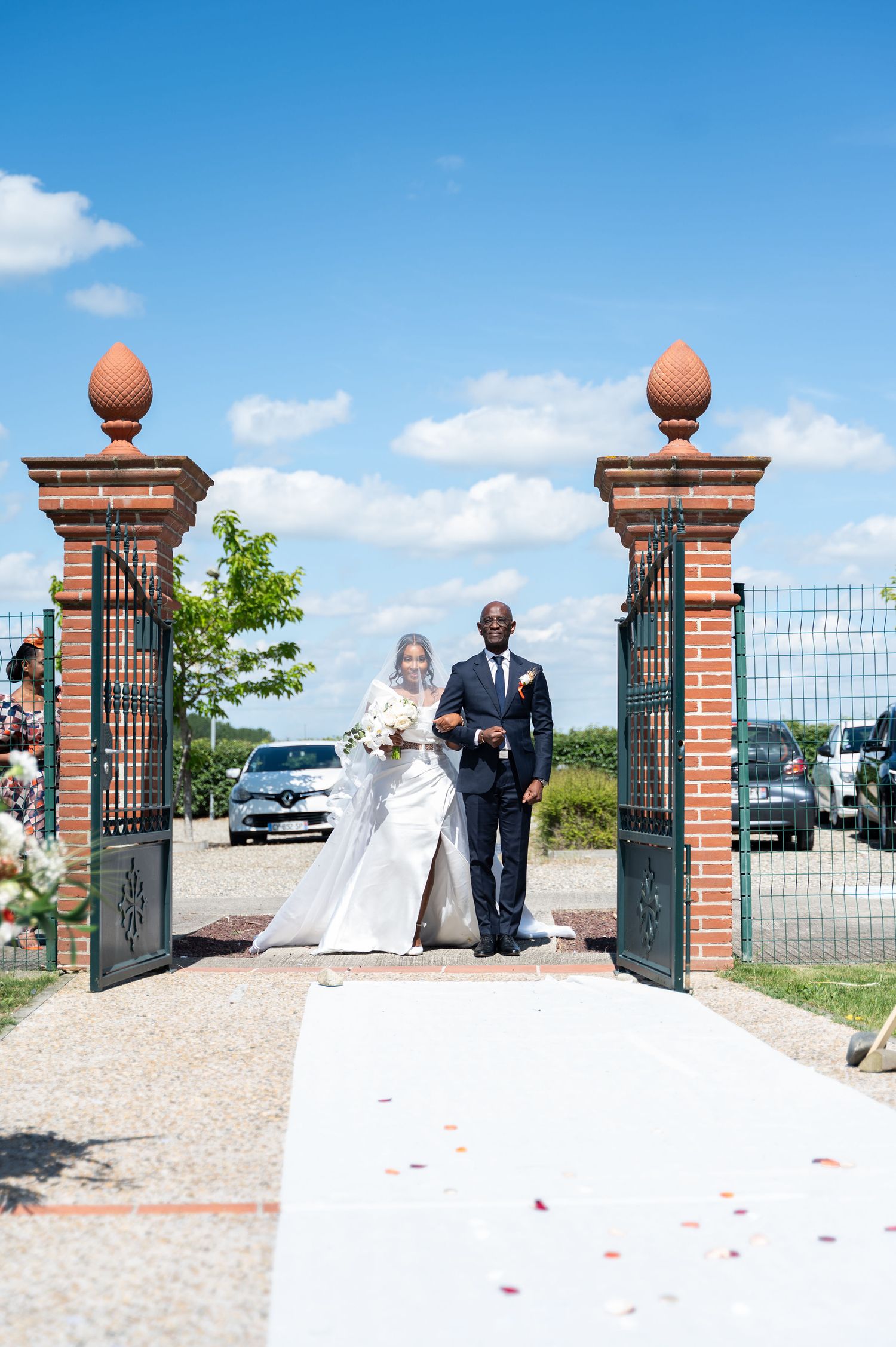 Bride in white gown walks through ornate iron gates with escort on sunny wedding day.