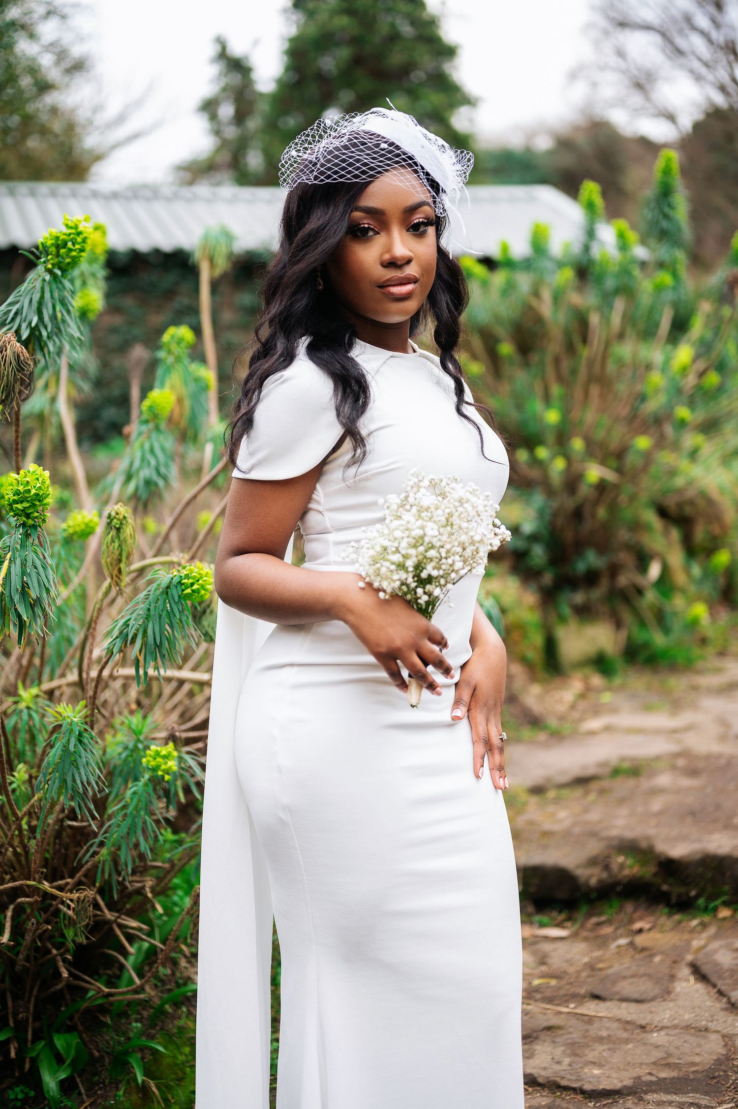 Elegant Black bride in white fitted gown holding baby's breath bouquet with birdcage veil in garden.