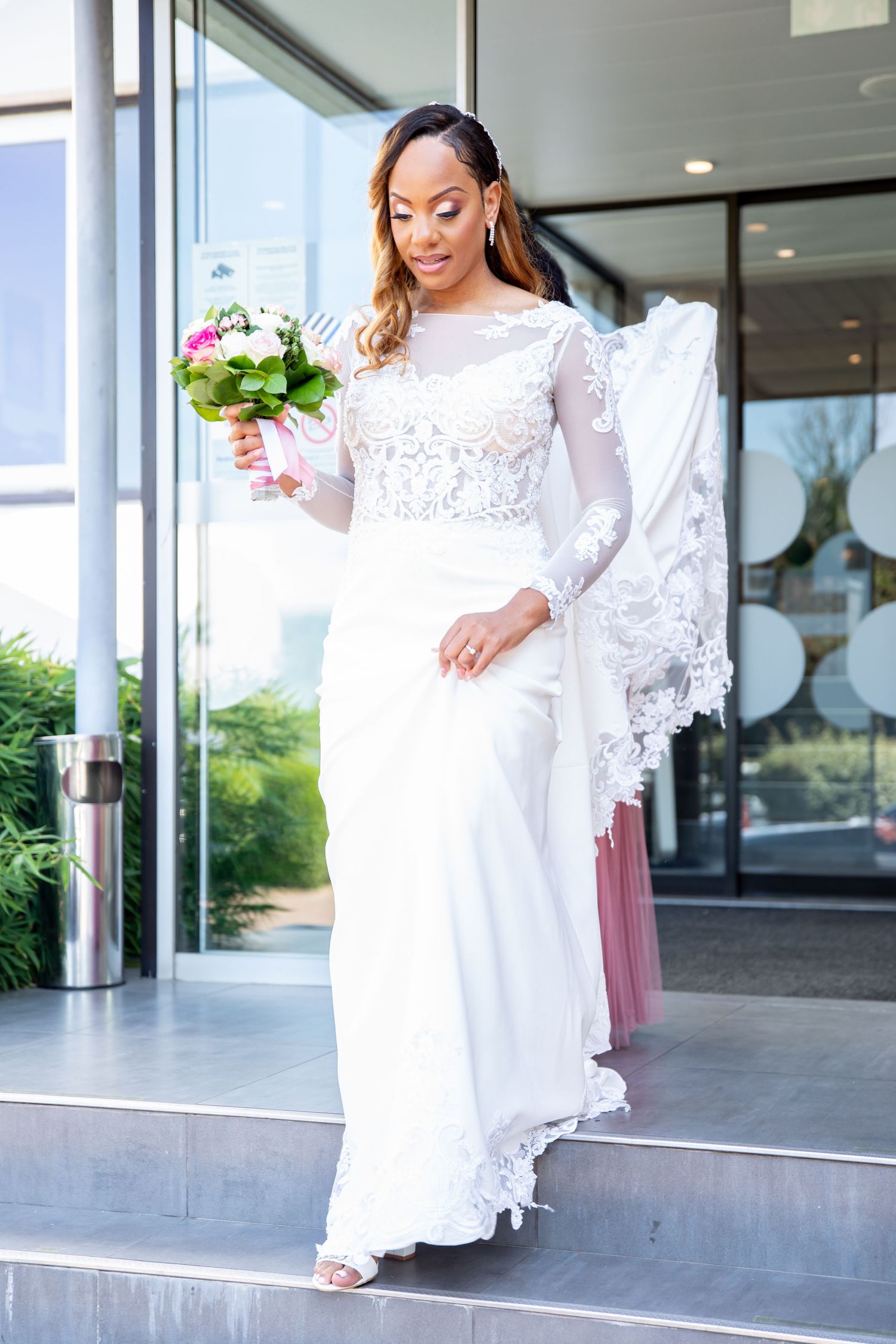 Elegant bride in white lace long-sleeve wedding gown holding a floral bouquet on building steps.