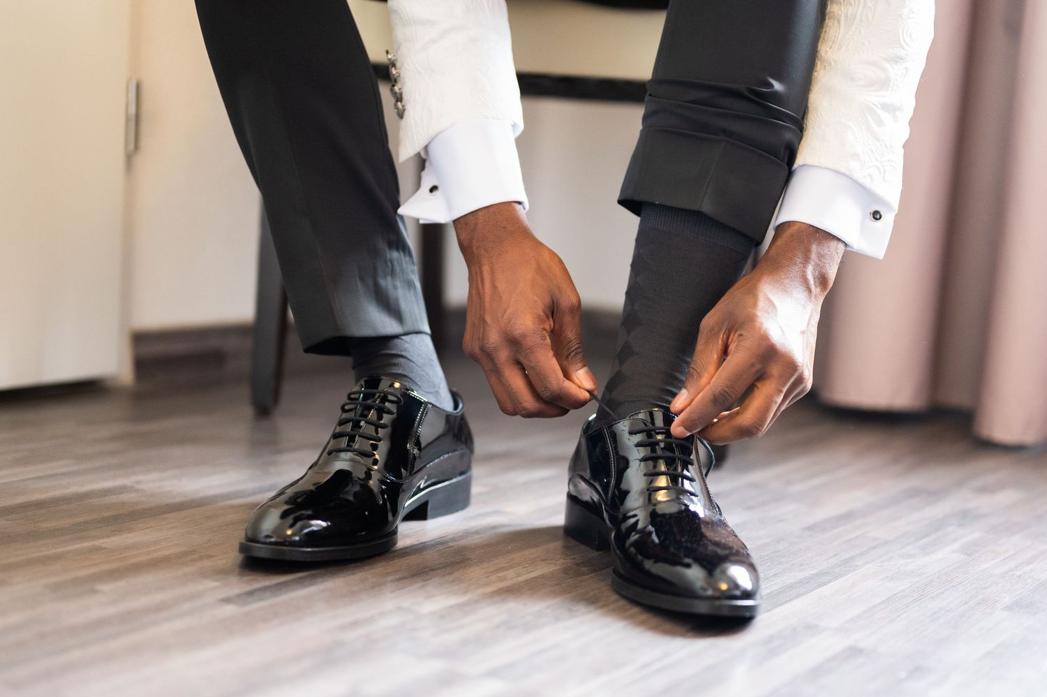 Man in dress shirt and gray trousers tying laces on shiny black patent leather Oxford shoes on hardwood floor.