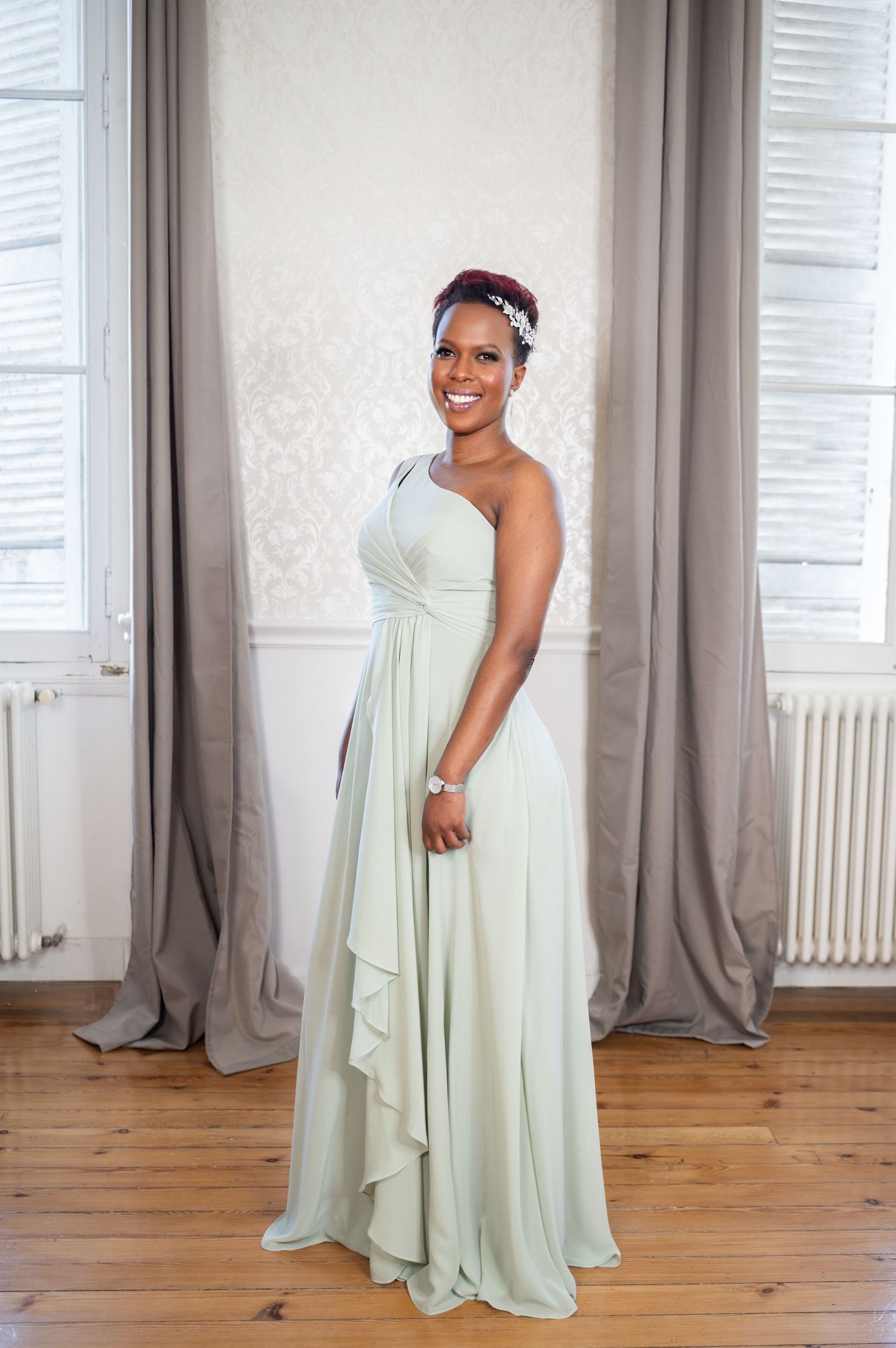 A smiling woman in an elegant sage green one-shoulder bridesmaid gown with a floral hair accessory.
