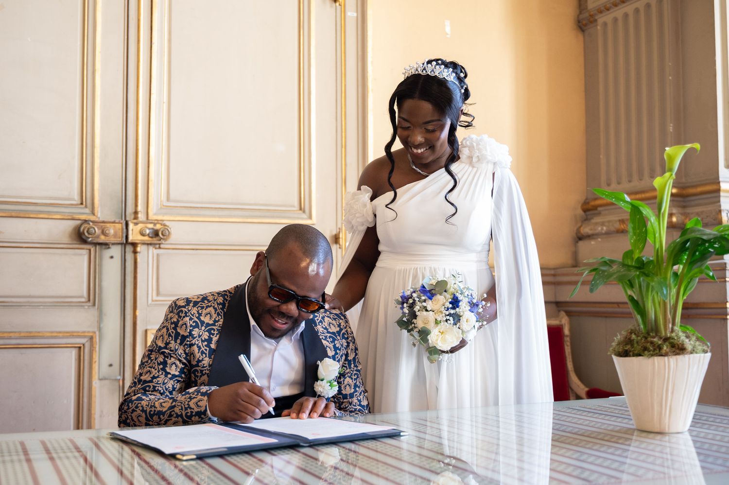 A groom in a floral suit signs the wedding registry while his smiling bride in white stands beside him holding flowers.