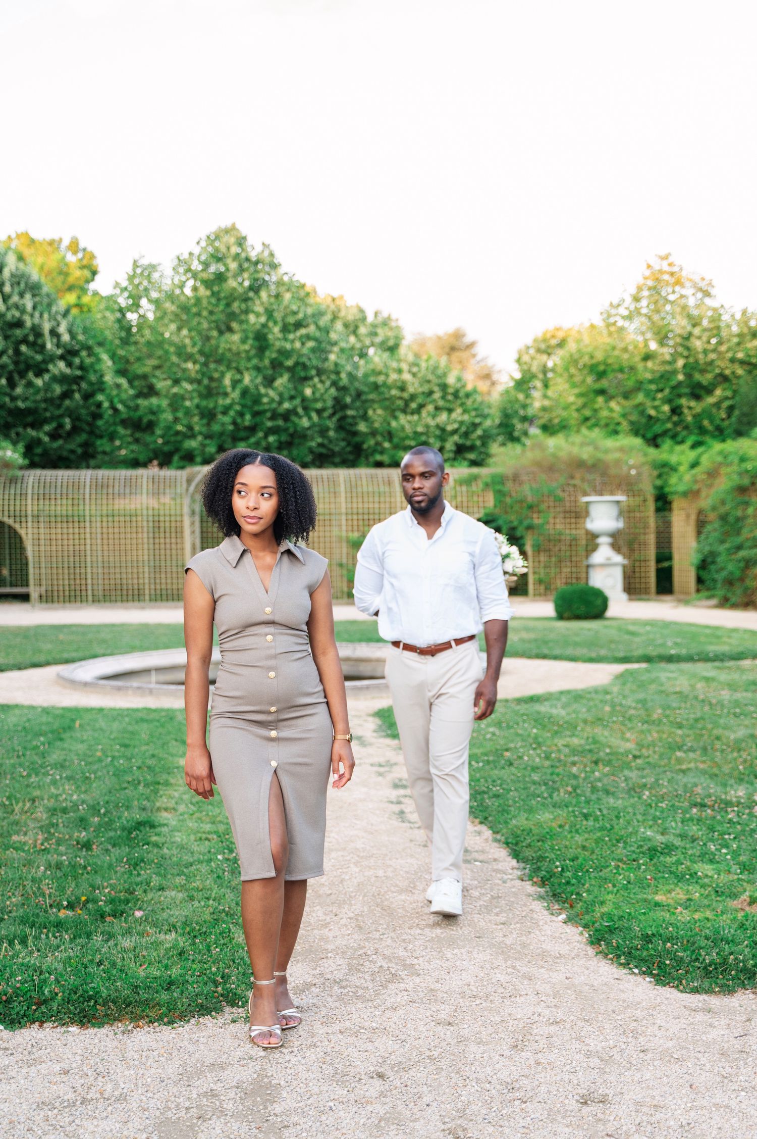A woman in a taupe dress walks confidently along a garden path as a man in white follows behind her.