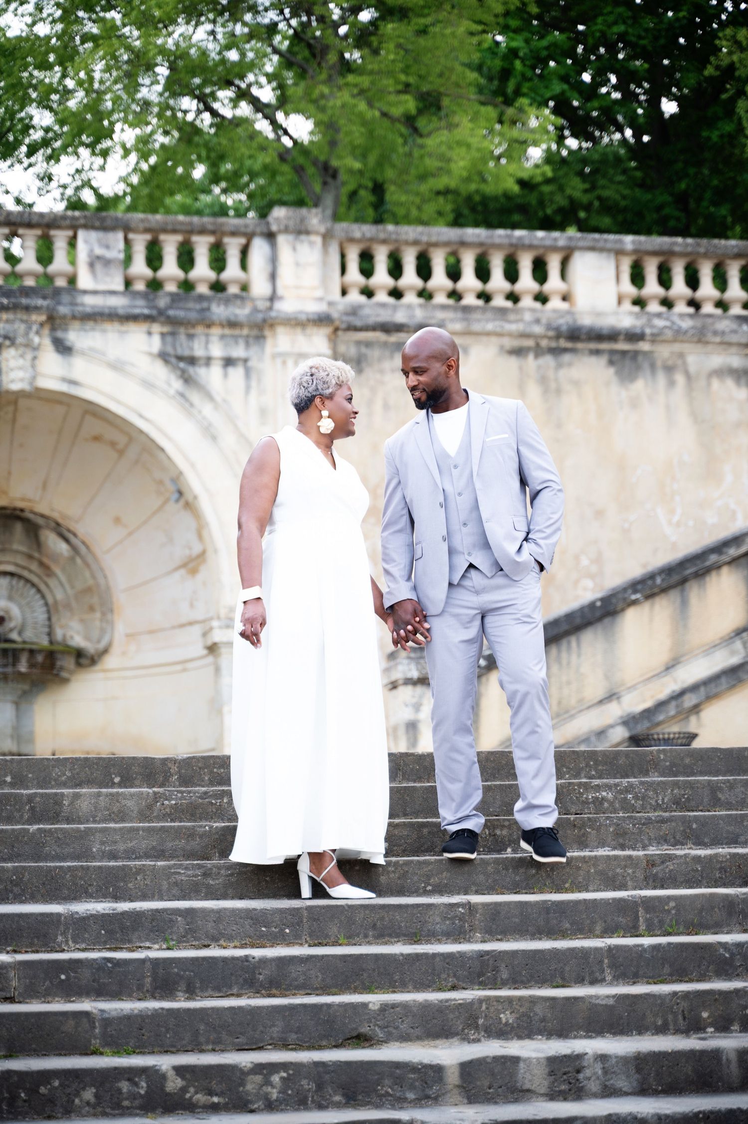 Black couple holding hands on stone staircase, woman in white dress and man in light gray suit.