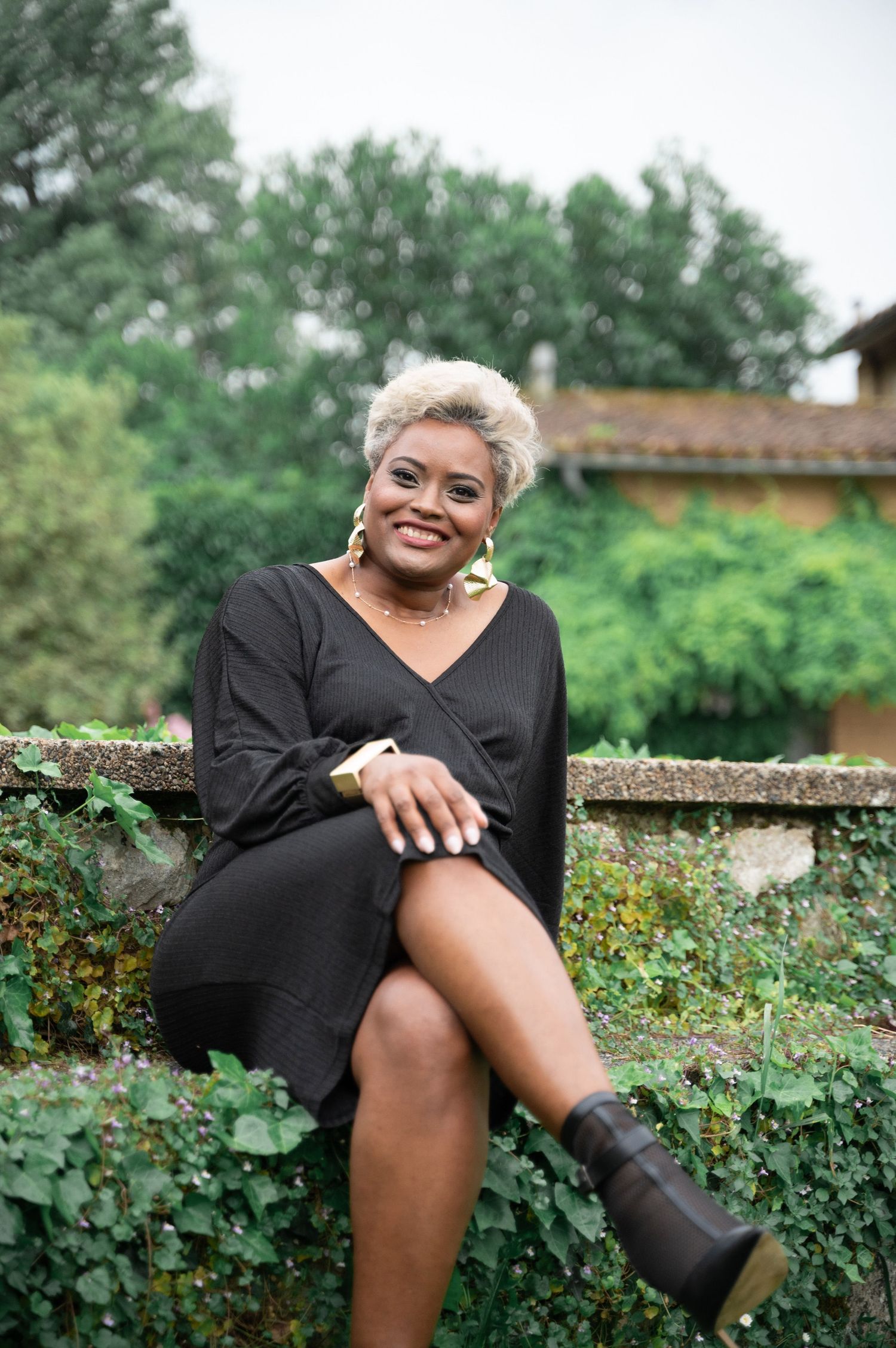 Smiling Black woman in black dress with gold earrings sitting on stone wall surrounded by green ivy.