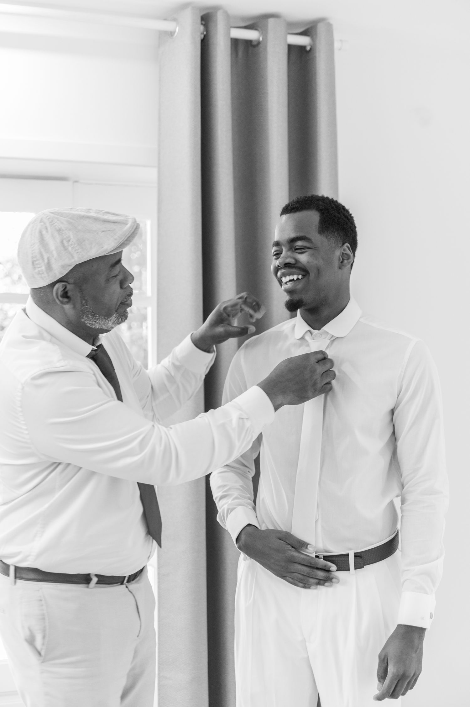 Black and white photo of older man helping young groom adjust his tie while both smile warmly.