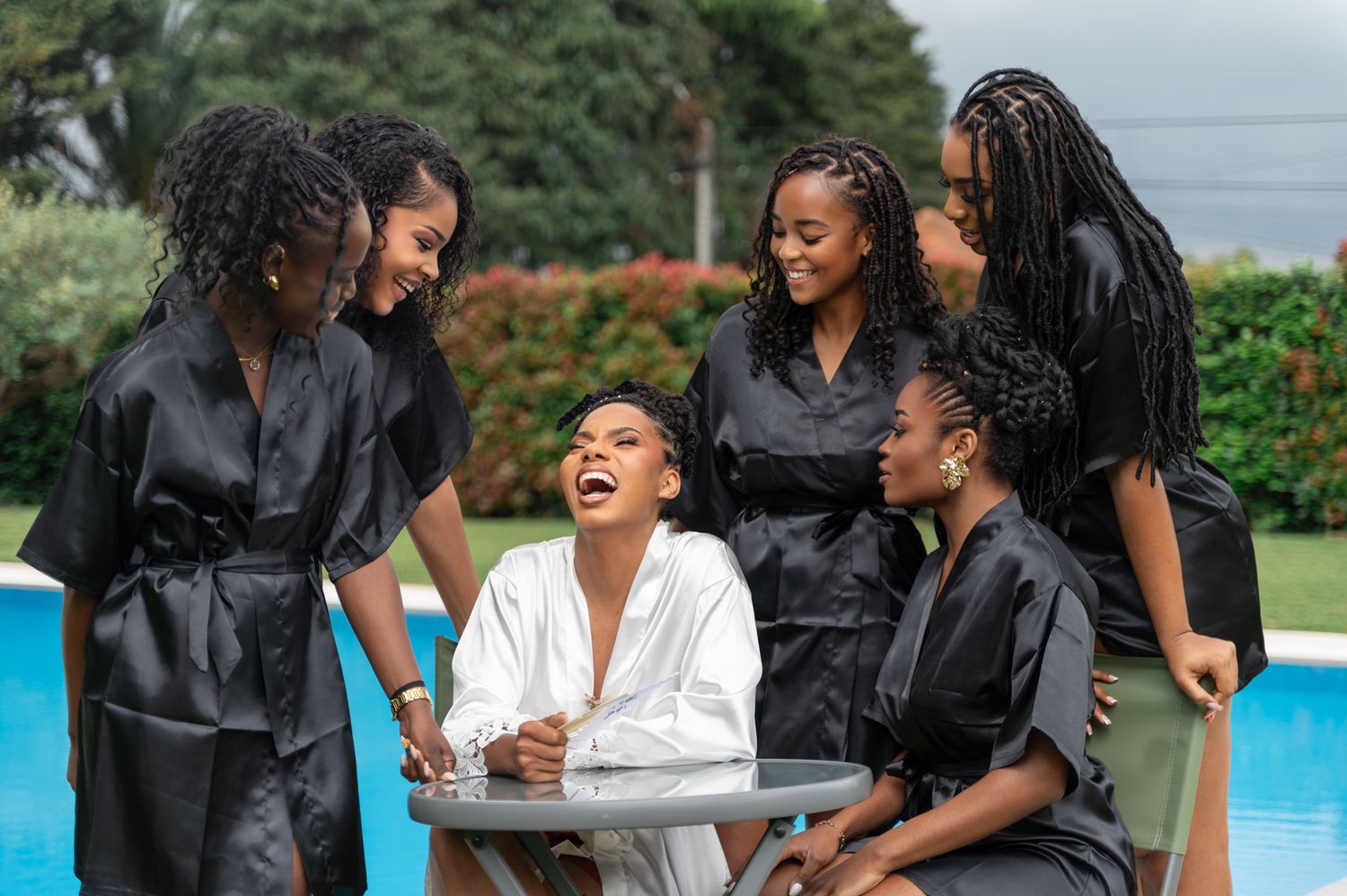 Bride in white robe laughing joyfully surrounded by bridesmaids in black robes by a pool.