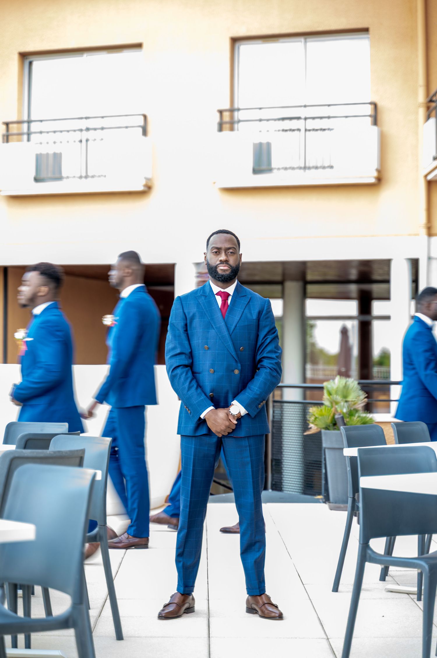 Confident Black groom in double-breasted blue suit with red tie poses outdoors while groomsmen walk behind.